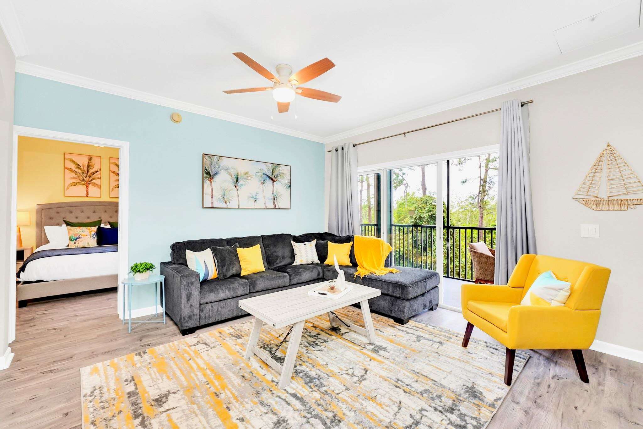 Living room with a gray sectional sofa, light blue accent wall, and a view into an adjacent bedroom through an open doorway, featuring a large palm tree artwork above the sofa.