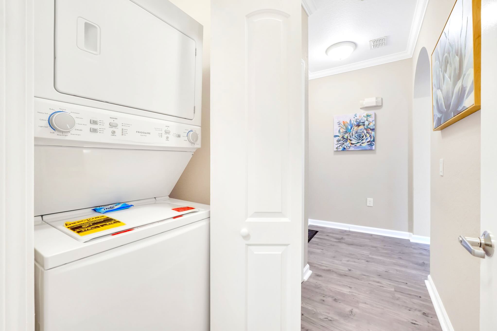 A white stacked washer and dryer unit is visible in a narrow hallway, with a partially open white door on the right leading to another room, featuring a small piece of paper on top of the washer.