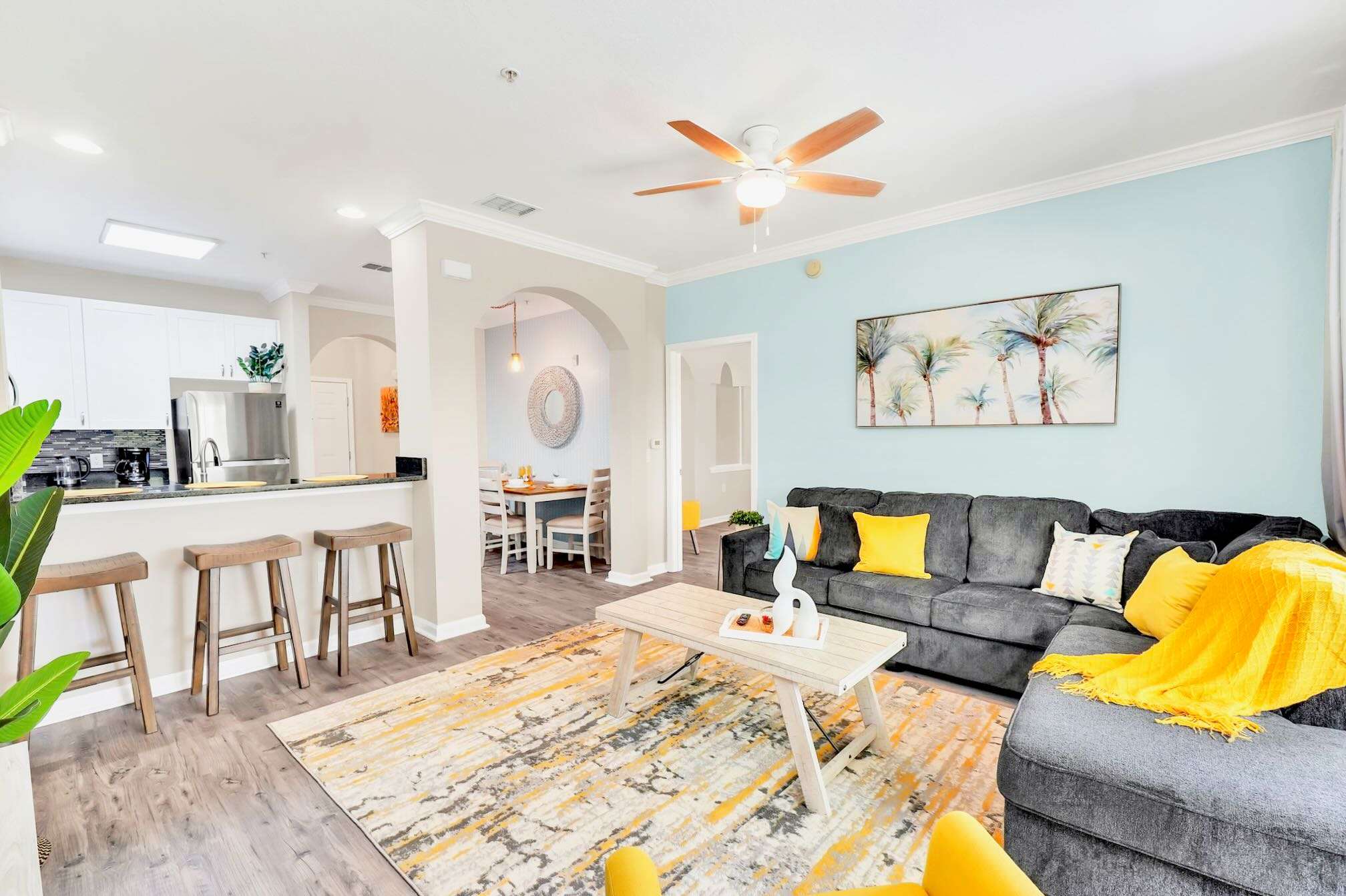Bright open-plan living room with a gray sectional sofa, light blue accent wall, and a view into the modern kitchen with white cabinetry and a breakfast bar with stools.