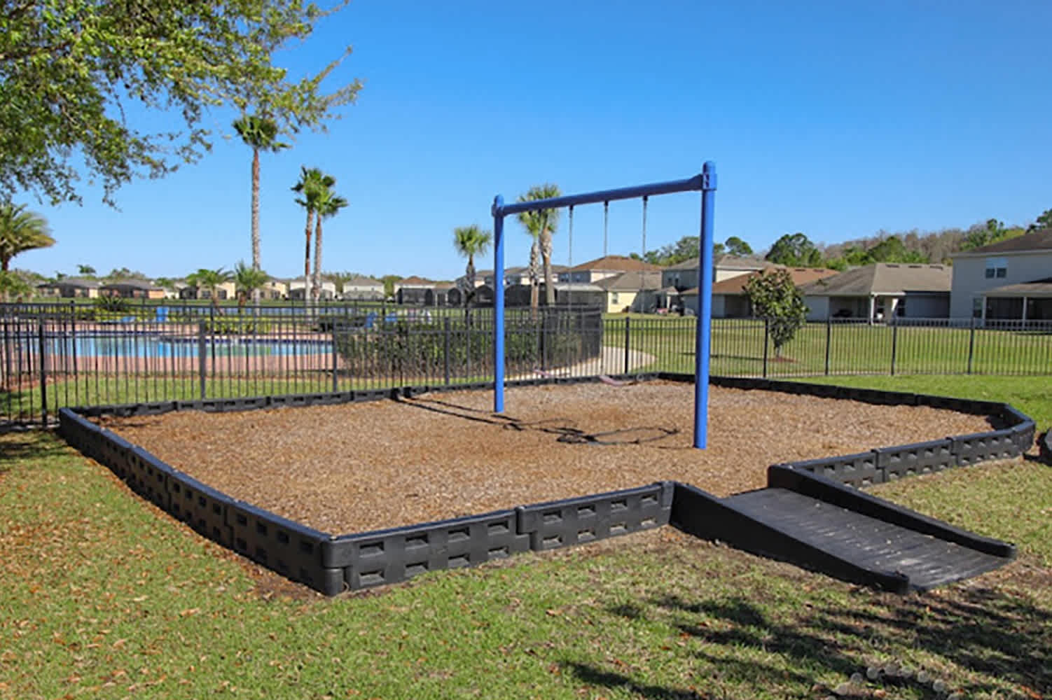 A blue metal swing set stands on a wood chip playground area, enclosed by a black plastic border with a ramp. Beyond a black fence, a communal pool and residential homes are visible under a clear blue sky, surrounded by green grass and palm trees.