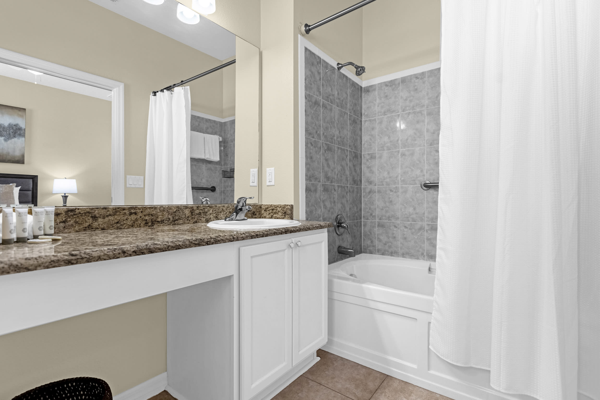 A functional bathroom with light beige walls, tiled flooring, and a granite-topped vanity with a single sink. A shower/tub combo with gray tiled walls and a white shower curtain is visible next to the vanity.