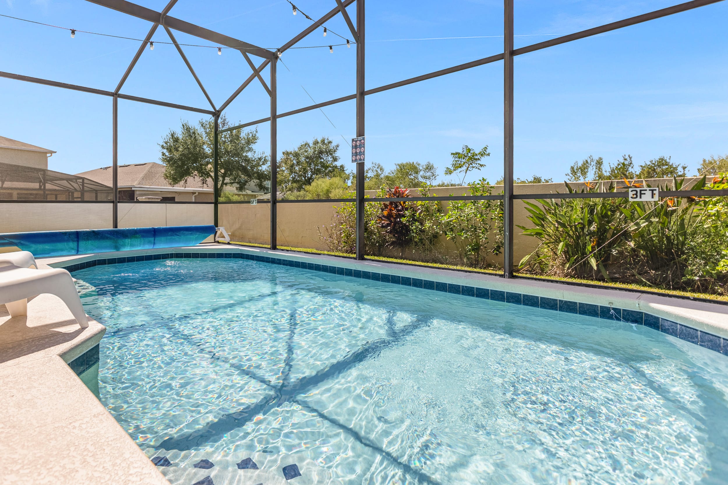 A sparkling blue private pool is enclosed within a screened lanai, featuring a shallow entry area and a pool cover rolled back. The surrounding patio is light-colored, with lush greenery visible outside the screen.