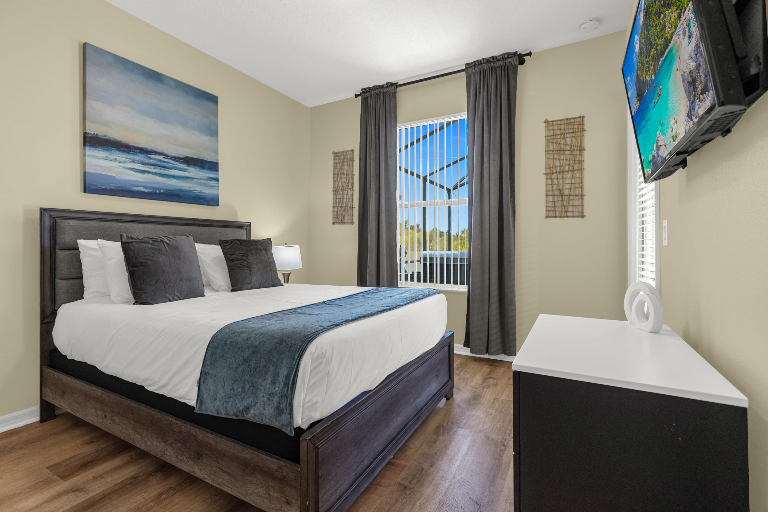 A modern bedroom with light beige walls, dark wood-look flooring, and a dark gray upholstered bed with white and blue bedding. A large window with vertical blinds and dark gray curtains overlooks an outdoor screened area, and a wall-mounted TV is visible.