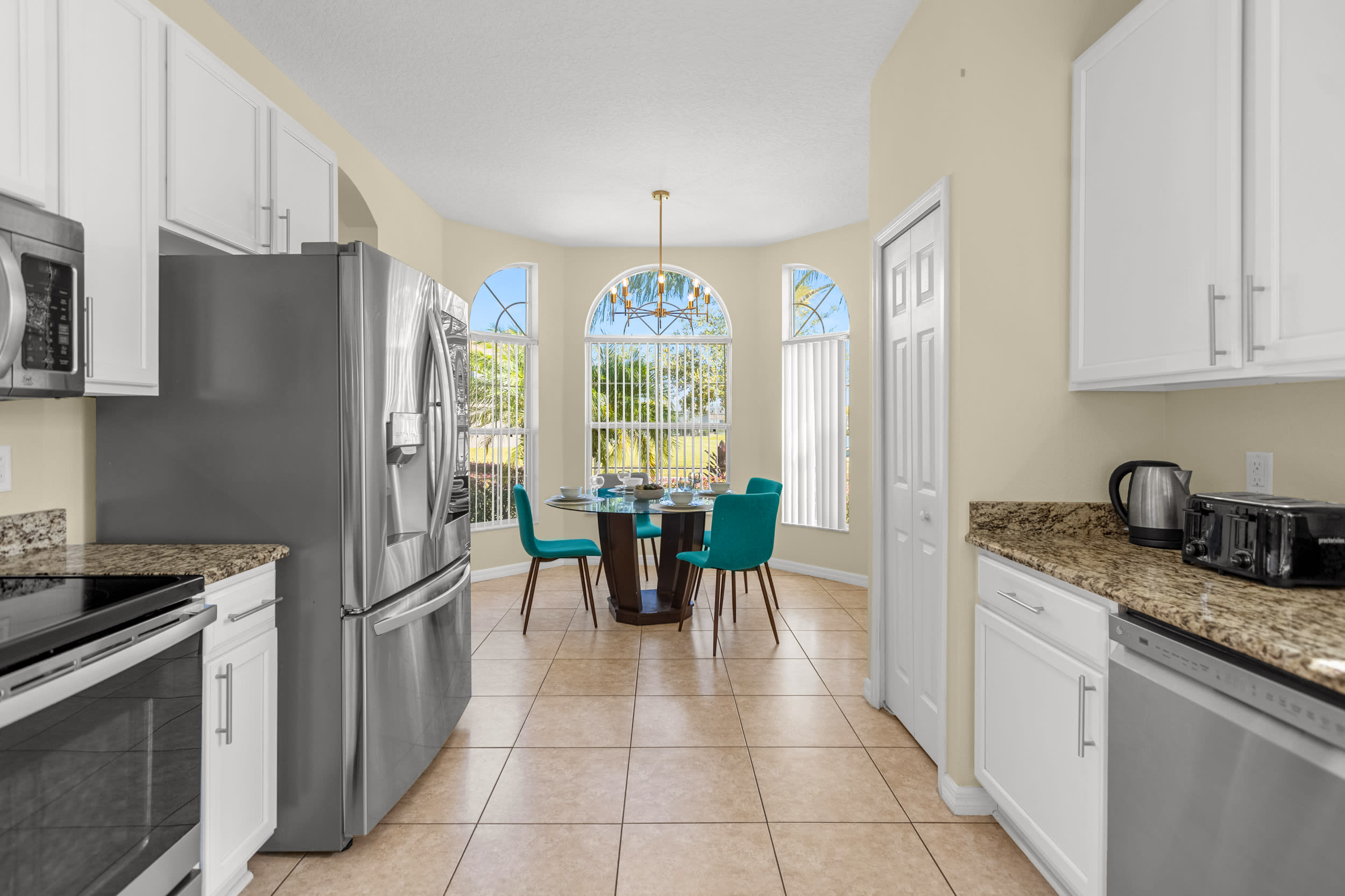 A view of the kitchen showcasing a large stainless steel refrigerator, black range, and white cabinets with granite countertops, leading to a bright breakfast nook with a round glass table and teal chairs.