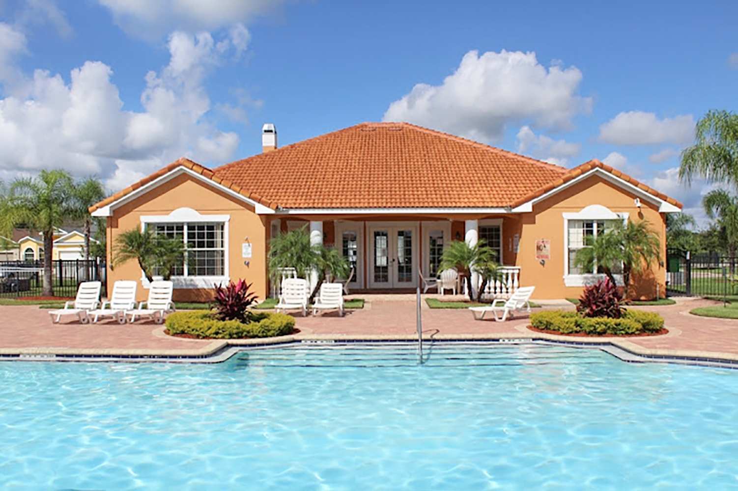 A vibrant orange stucco clubhouse with a red tile roof stands behind a large, clear blue communal swimming pool. White lounge chairs are arranged on the brick-paved deck, surrounded by lush green landscaping and palm trees under a bright blue sky.