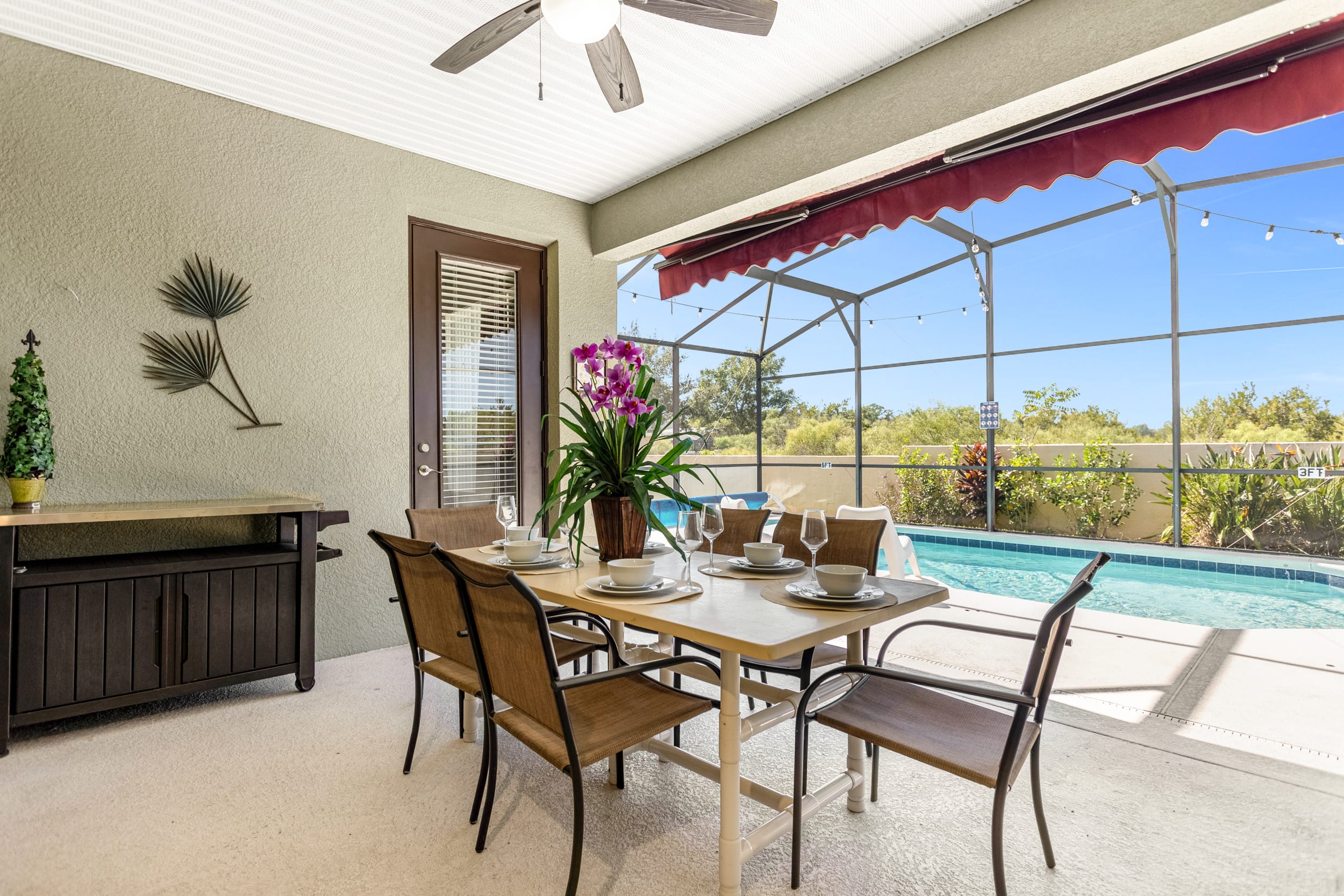 A covered outdoor patio features a dining table set for six with brown chairs, adorned with a vibrant orchid centerpiece. A red retractable awning provides shade, and a glass door leads indoors, with the screened pool visible beyond.