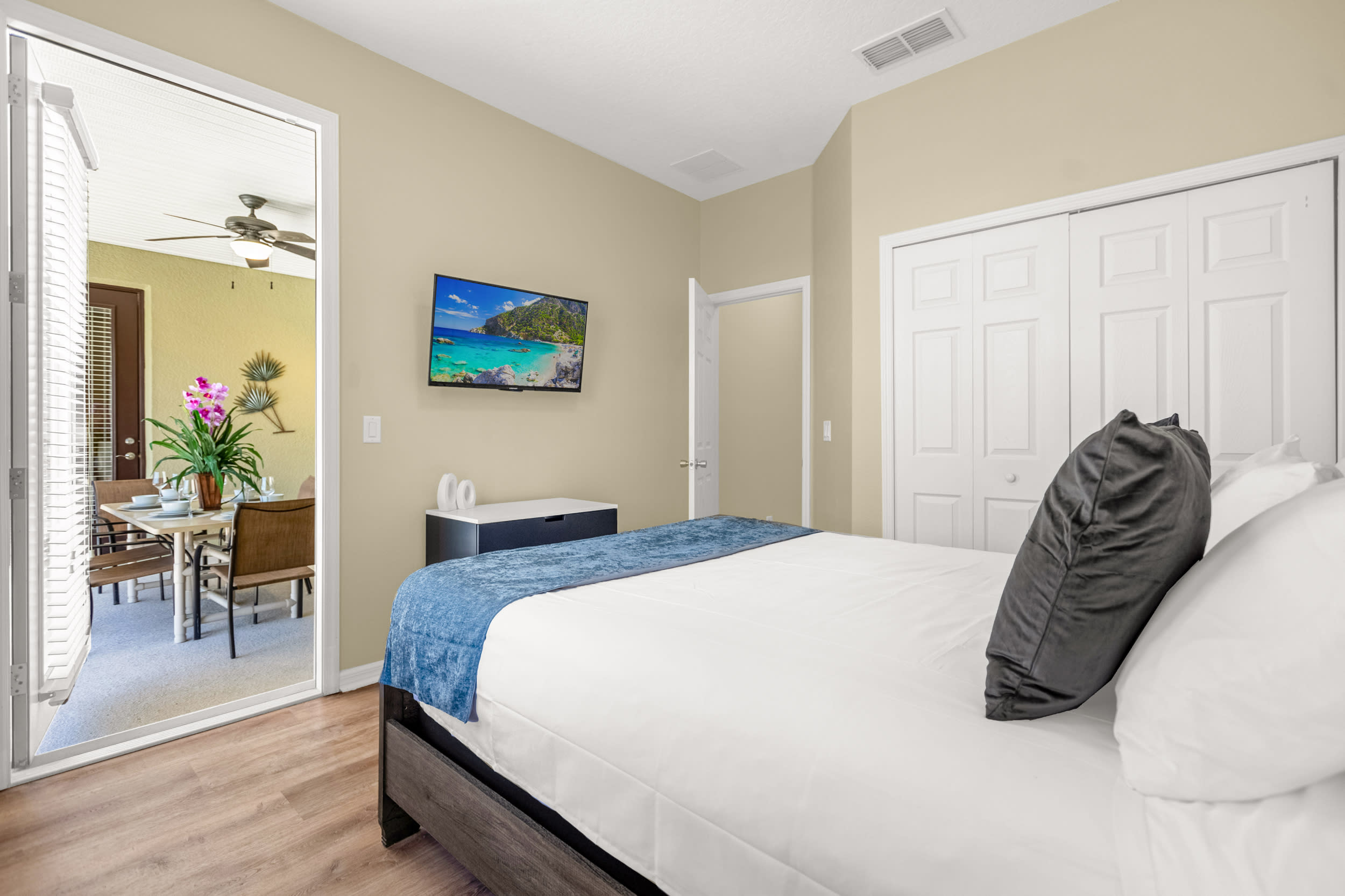A bright bedroom featuring a dark wood bed with white and blue bedding, light beige walls, and wood-look flooring. A wall-mounted TV is visible, and a sliding glass door opens to an outdoor patio with dining furniture.