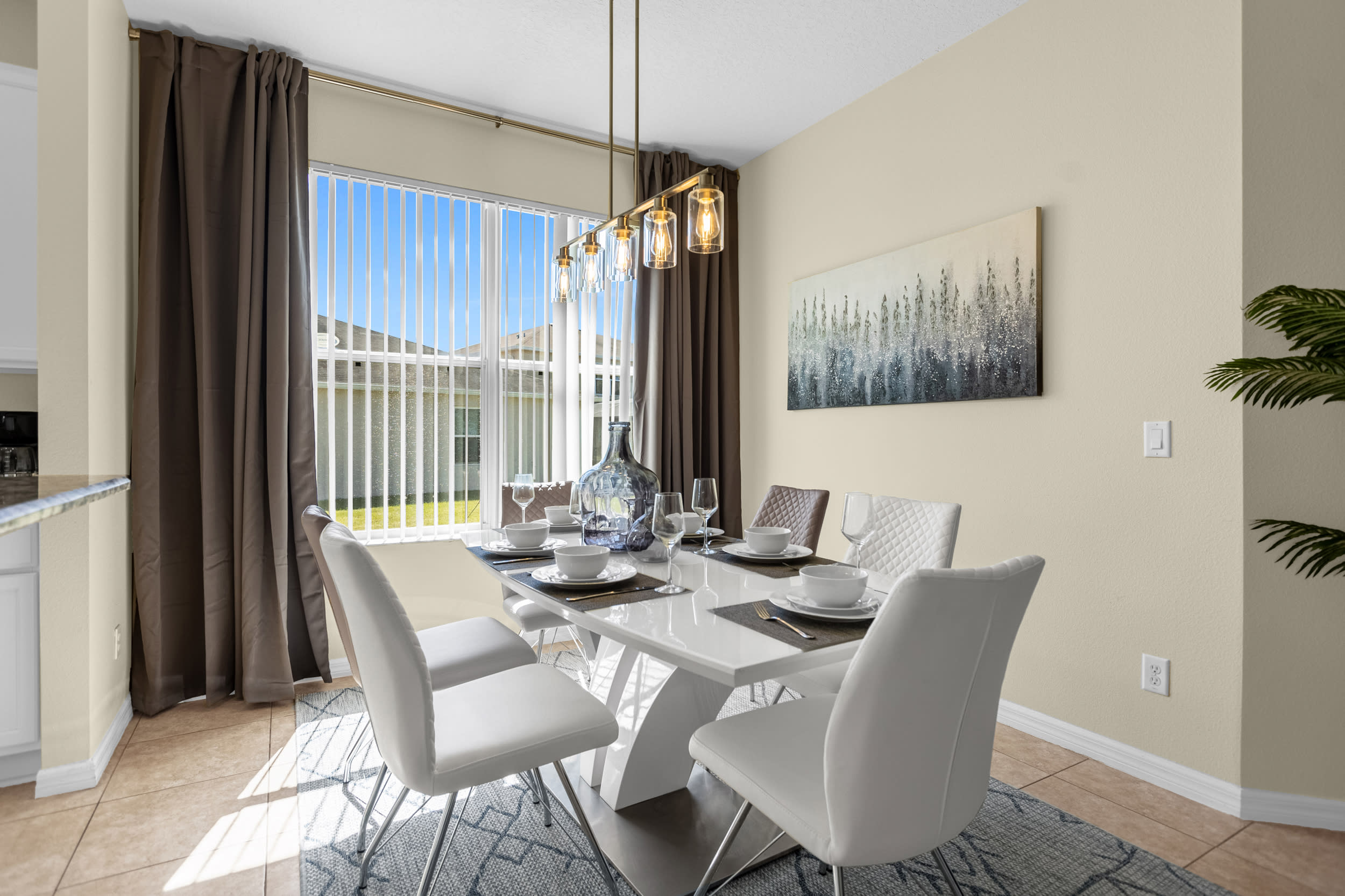 A bright dining area features a modern white rectangular table with six white and brown chairs, set on a patterned rug. A contemporary linear chandelier hangs above, and a large window with vertical blinds offers natural light, framed by dark brown curtains.