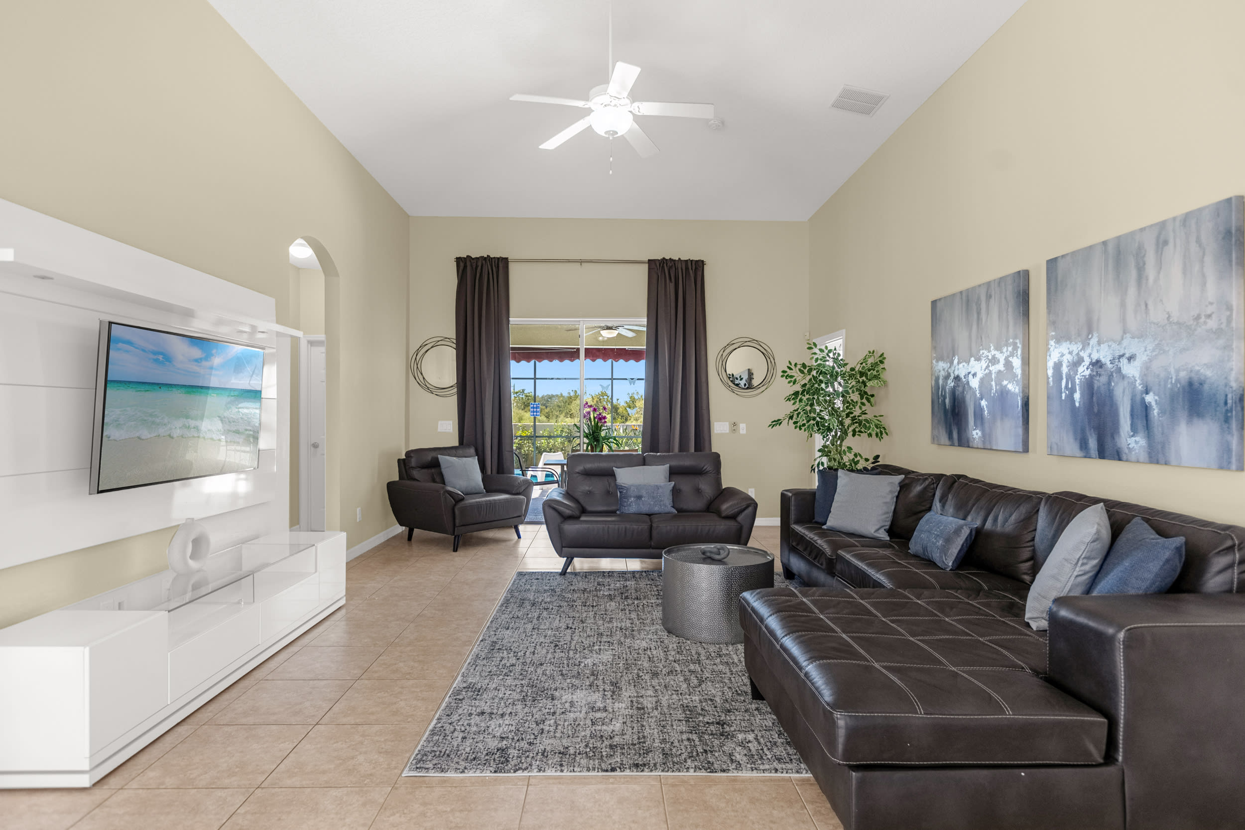 A spacious living room features a large dark brown sectional sofa and a matching armchair, centered around a round metallic coffee table on a gray patterned rug. A large wall-mounted TV on a white entertainment unit and abstract artwork adorn the light beige walls.
