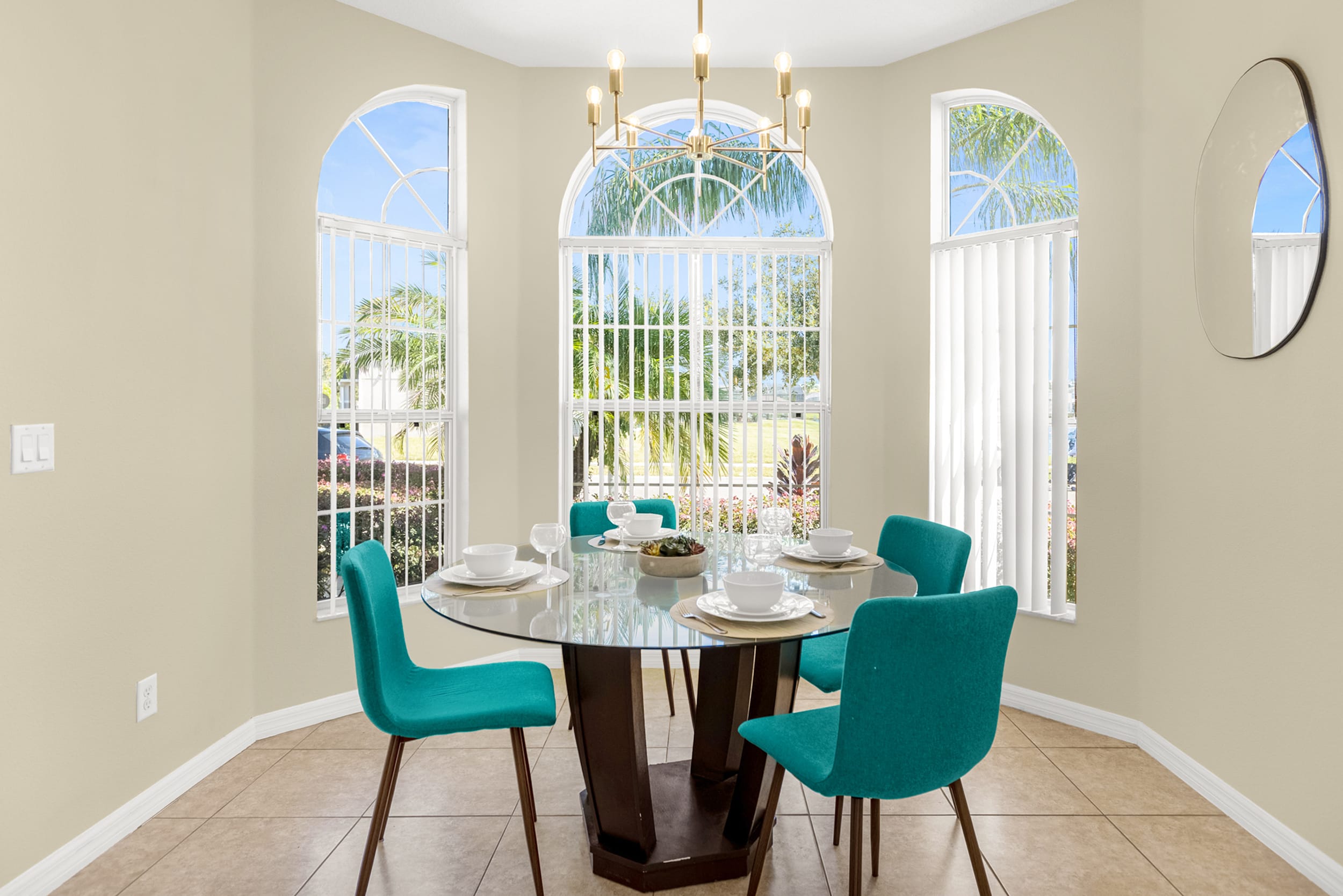 A sunlit breakfast nook featuring a round glass table with teal chairs, positioned in a bay window area with arched windows and vertical blinds, illuminated by a modern gold chandelier.