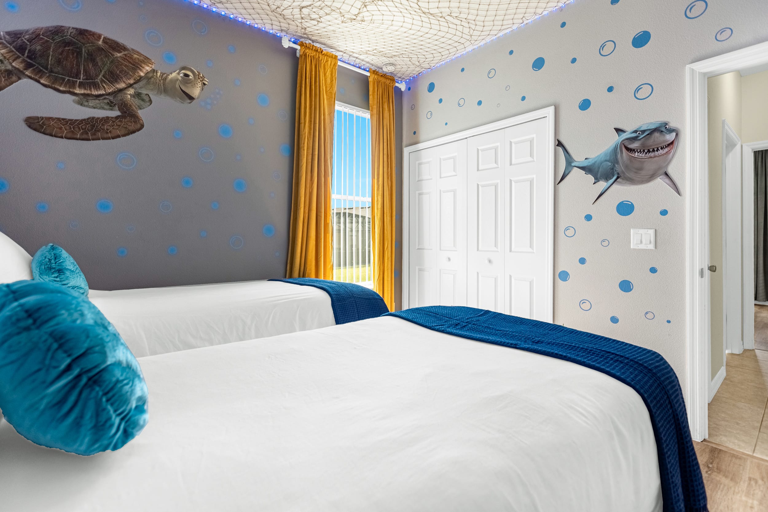 A fun, themed bedroom with two twin beds and wood-look flooring, featuring a gray wall adorned with blue bubbles and a large sea turtle and shark decor. Yellow curtains frame a window, and a decorative fishing net covers the ceiling.