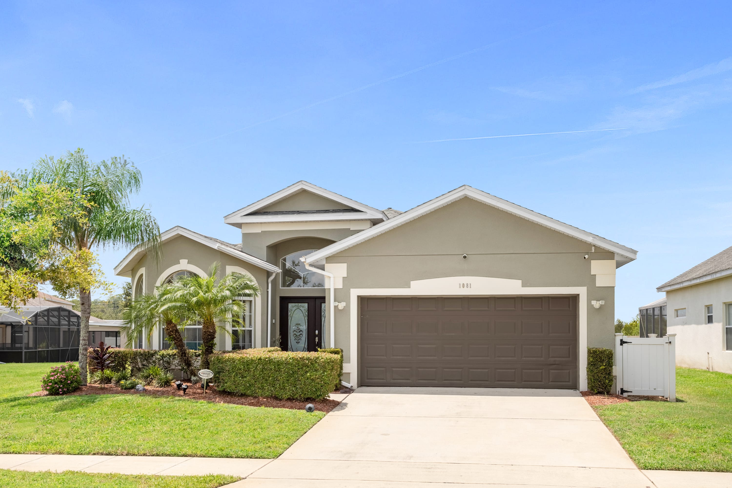 A modern single-story house with a light gray stucco exterior, a dark brown garage door, and a well-maintained green lawn. Palm trees and shrubs decorate the front, and a concrete driveway leads to the two-car garage.