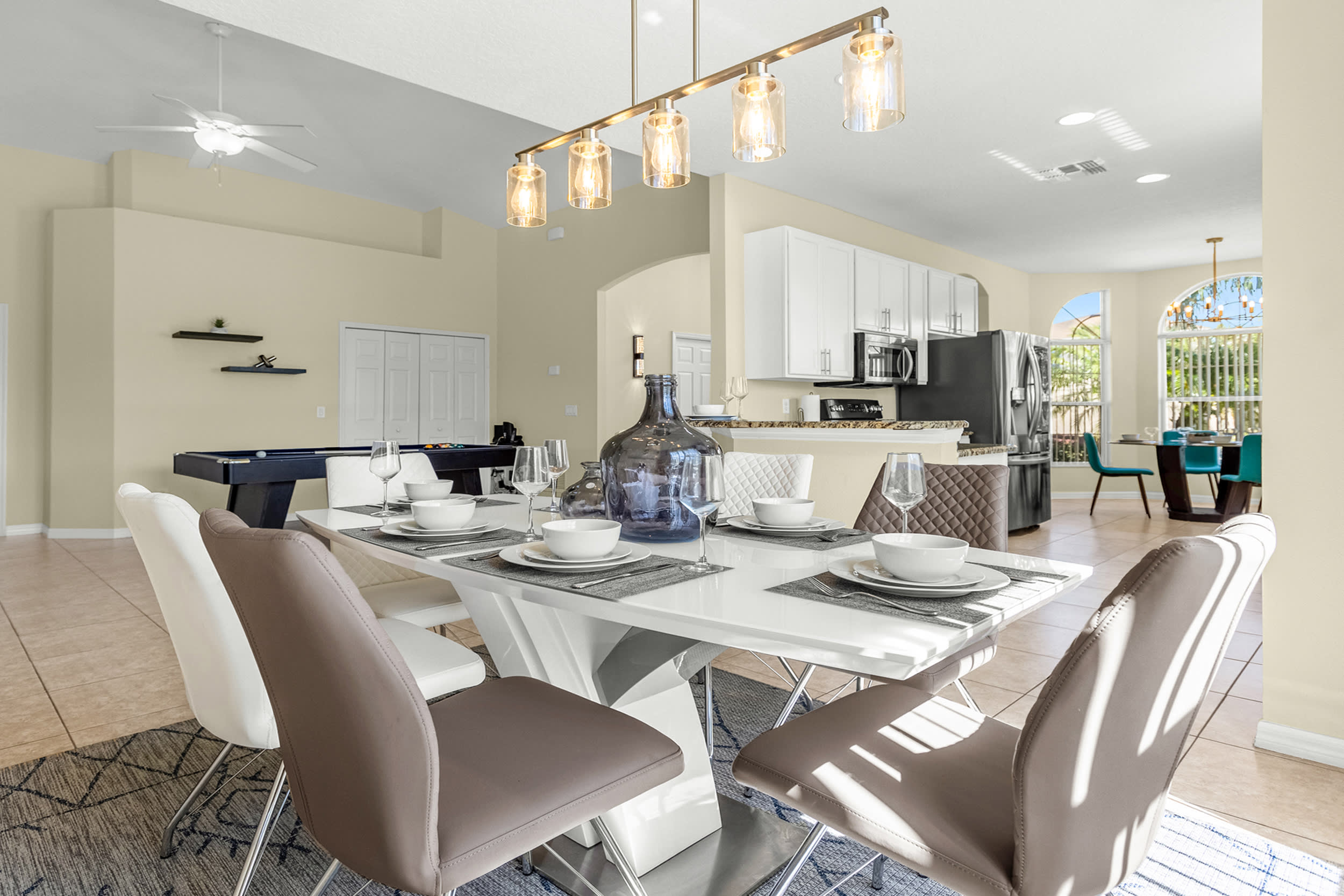 A modern dining area features a white rectangular table set with white dishes and a large blue glass vase, surrounded by white and brown upholstered chairs, with a pool table visible in the background.