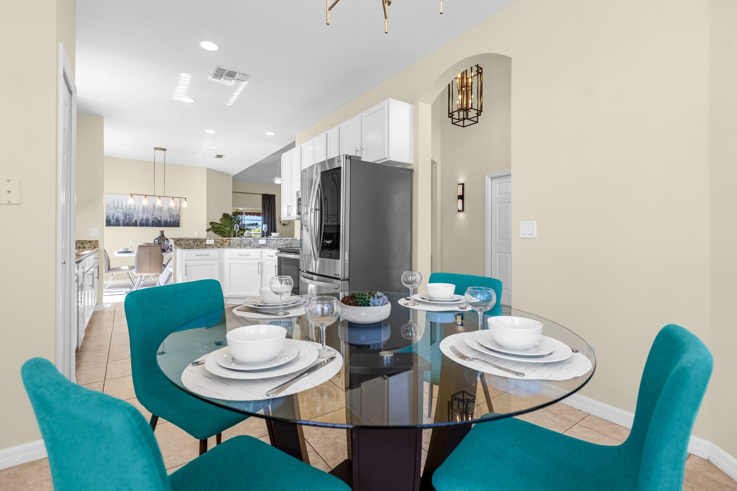 A cozy breakfast nook with a round glass-top table set with white dishes and a small plant, surrounded by four vibrant teal upholstered chairs, with the kitchen visible in the background.