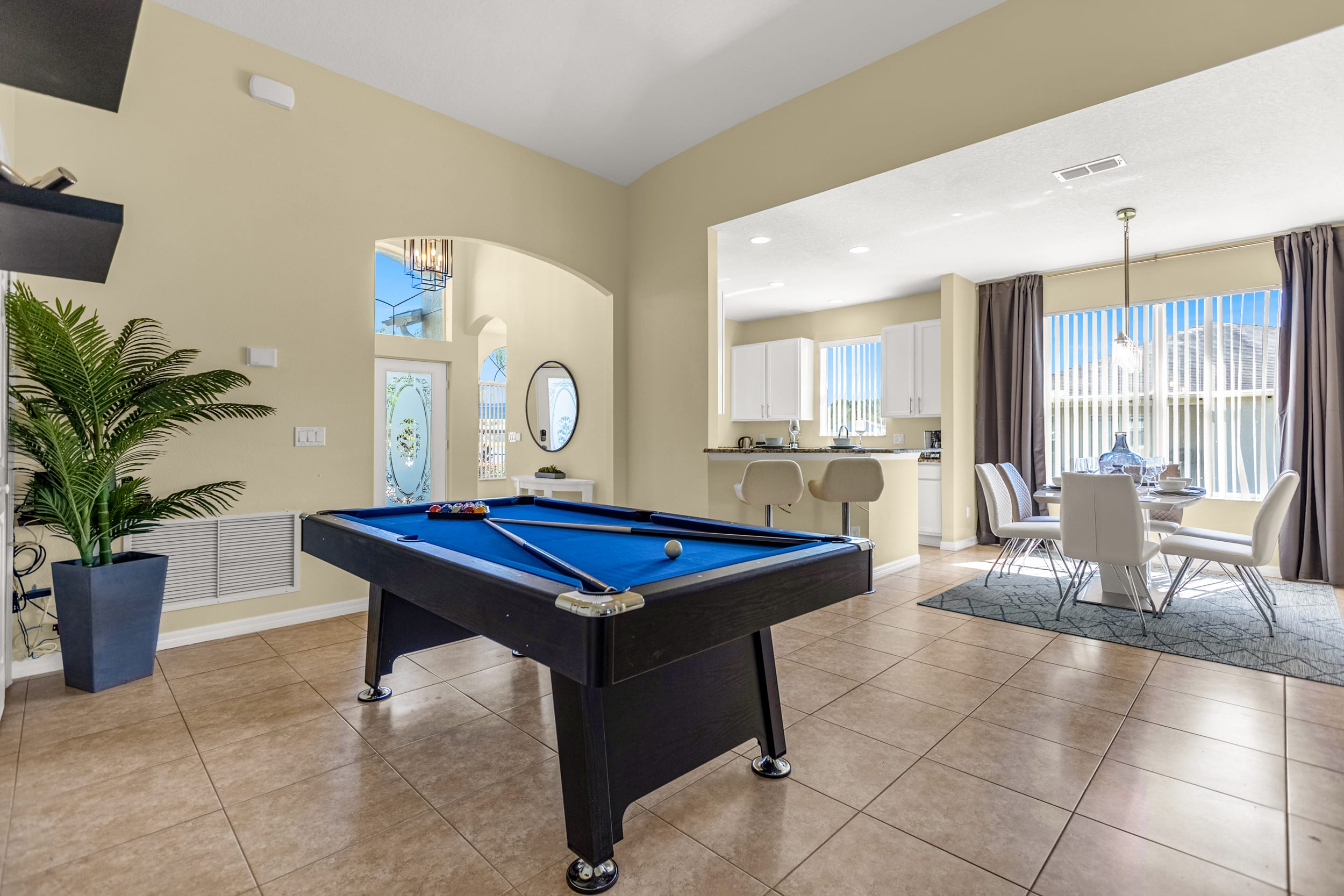 A wide shot of the game room shows a blue-felted pool table as the central feature, with an open view towards the front entrance foyer and a glimpse of the kitchen and dining area in the background. The room has light beige walls and tiled flooring.