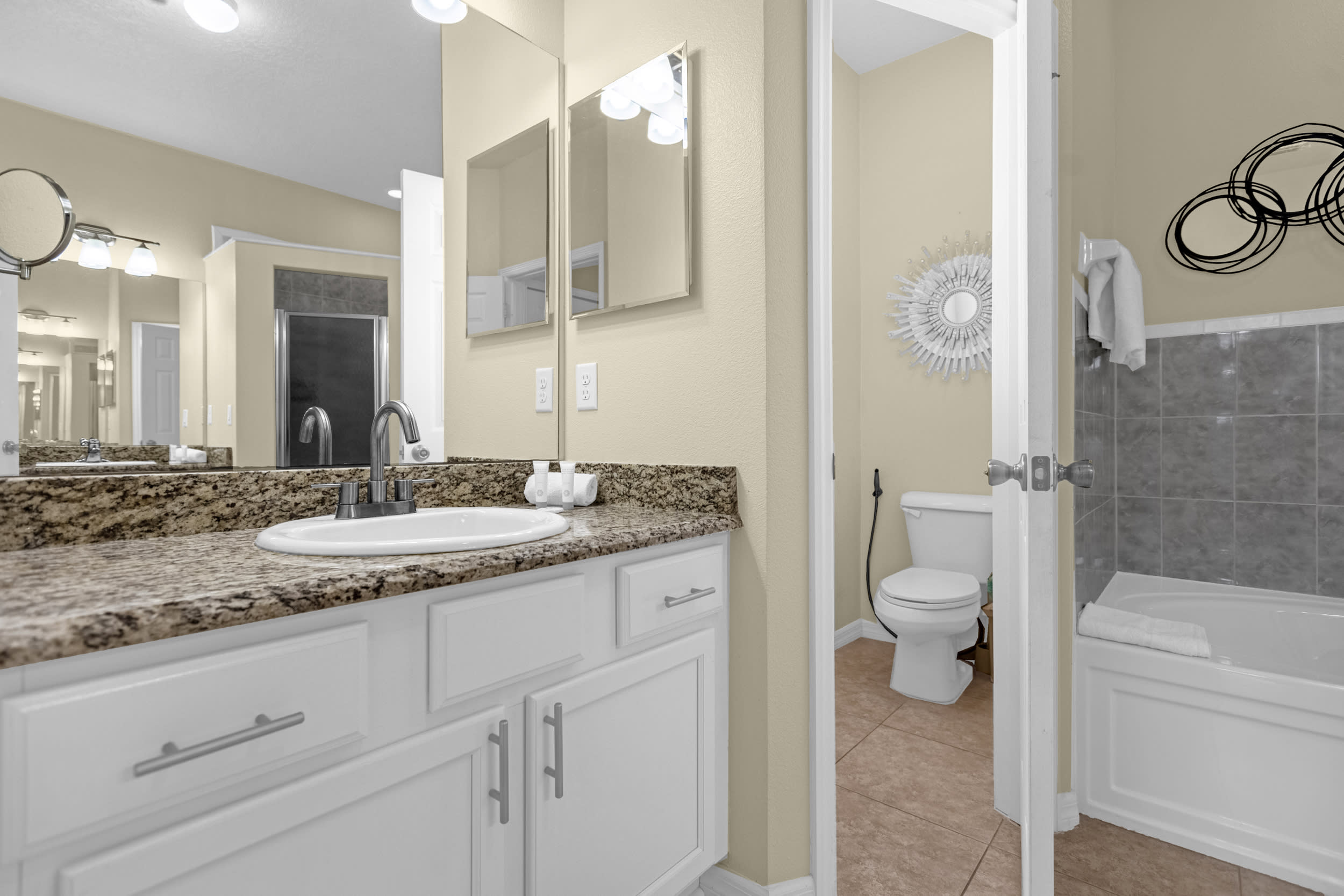 A bathroom vanity with a single sink, granite countertop, and white cabinets, reflecting a mirror, with a toilet visible in an adjacent alcove and a bathtub in the background.