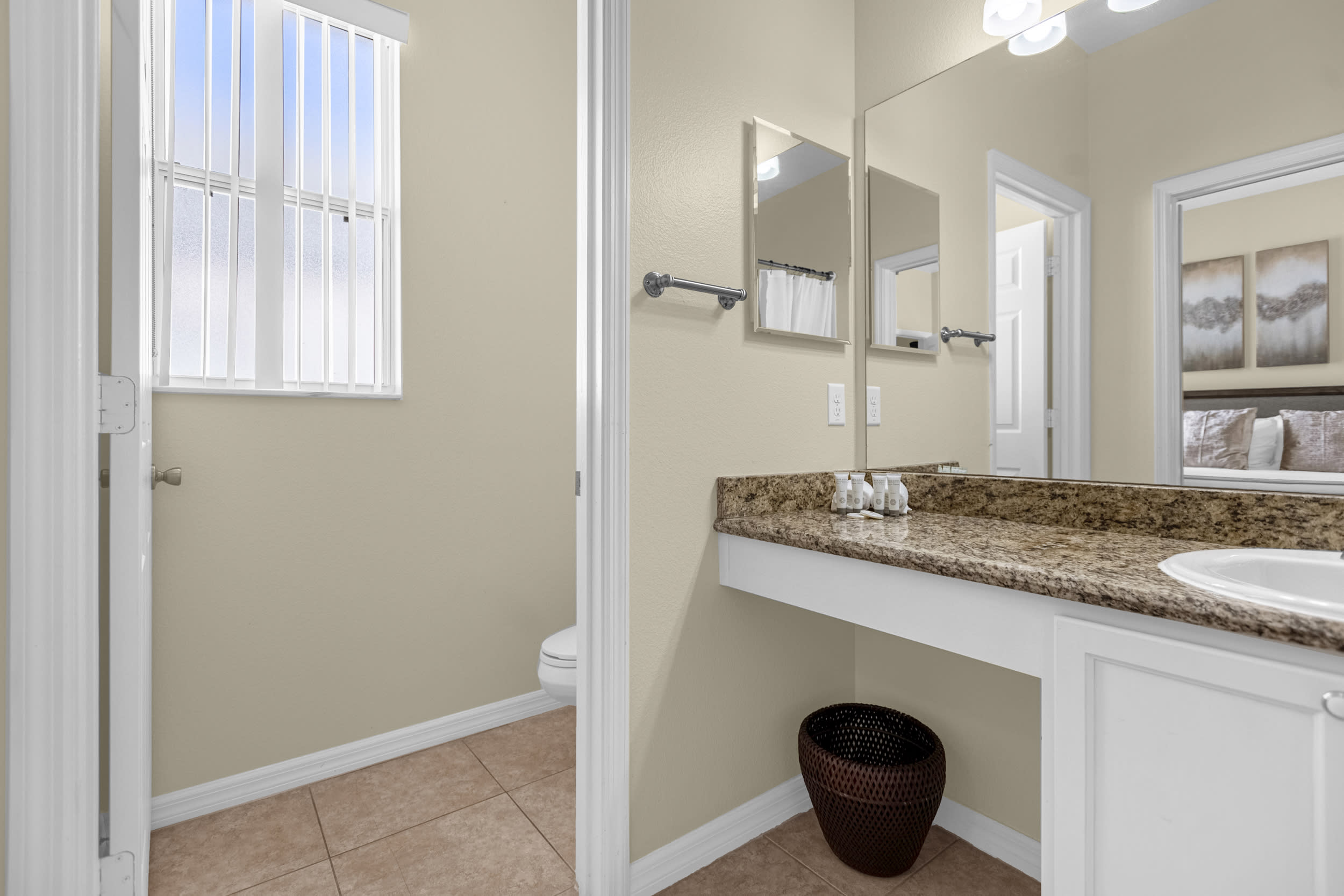 A bright bathroom with light beige walls and tiled flooring, featuring a granite-topped vanity with a single sink and a separate toilet area with a window. The room is well-lit and includes a large mirror above the vanity.