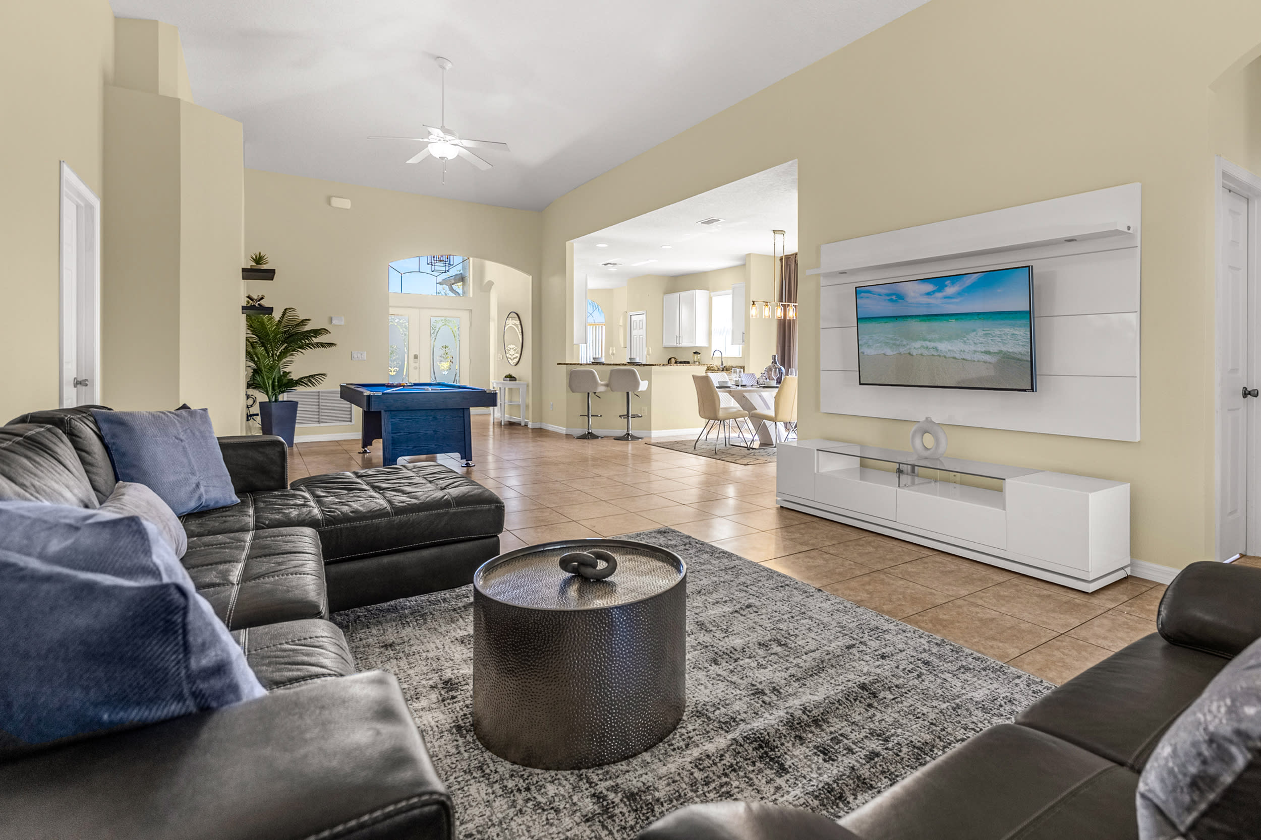 An expansive open-concept living area features a large dark leather sectional sofa facing a modern white entertainment unit with a wall-mounted TV. The space flows into a game room with a pool table and a dining area, all with tiled floors and light beige walls.