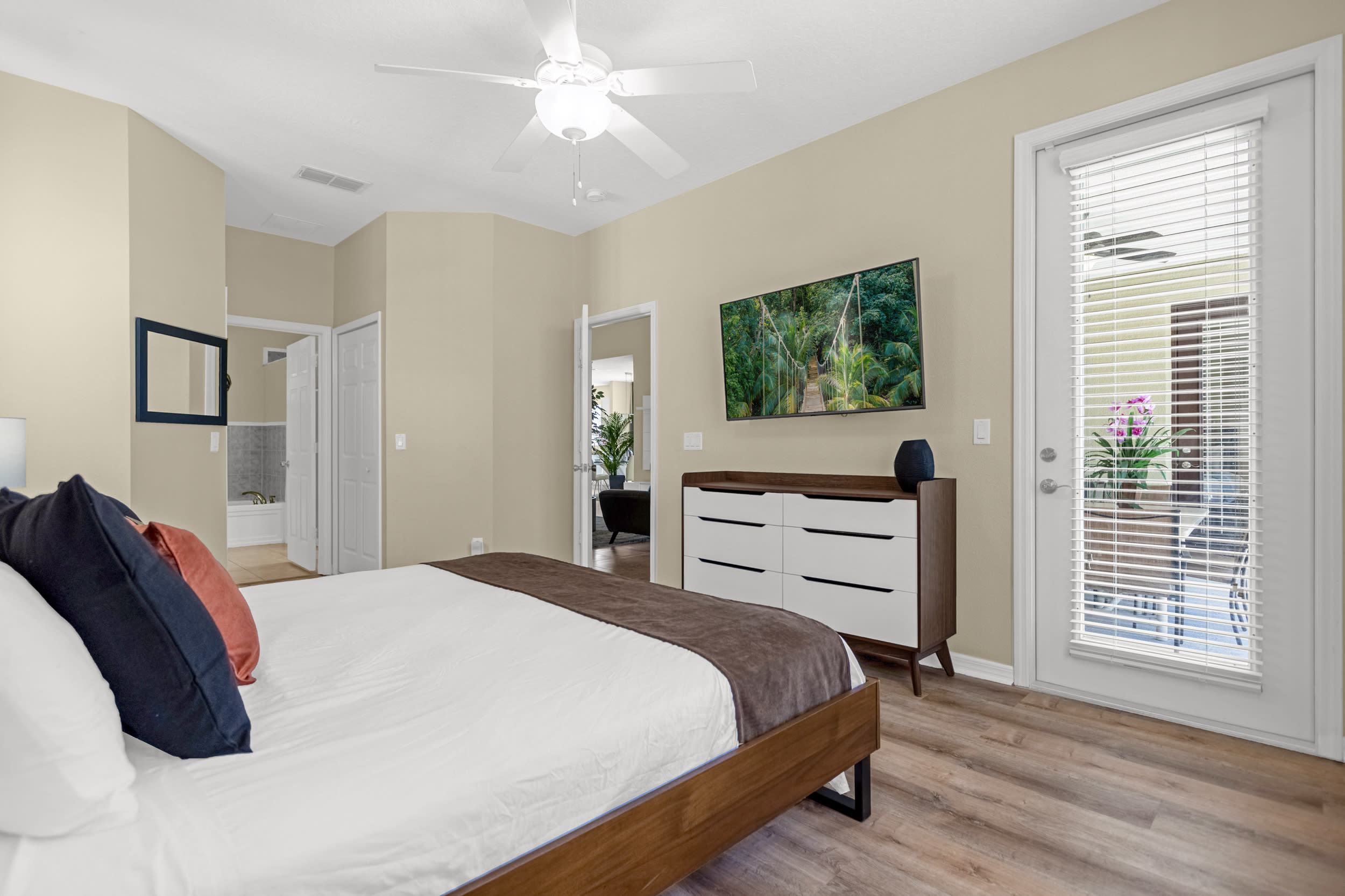 A spacious bedroom with a queen-sized bed featuring a wooden frame and white bedding, a modern dresser with a TV mounted above it, and a glass door leading to an outdoor area.