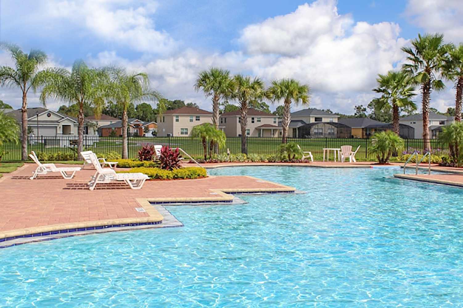 A large, irregularly shaped communal swimming pool with clear blue water is surrounded by a brick-paved deck and white lounge chairs. Palm trees and lush greenery line the fence, with residential homes visible in the sunny background.