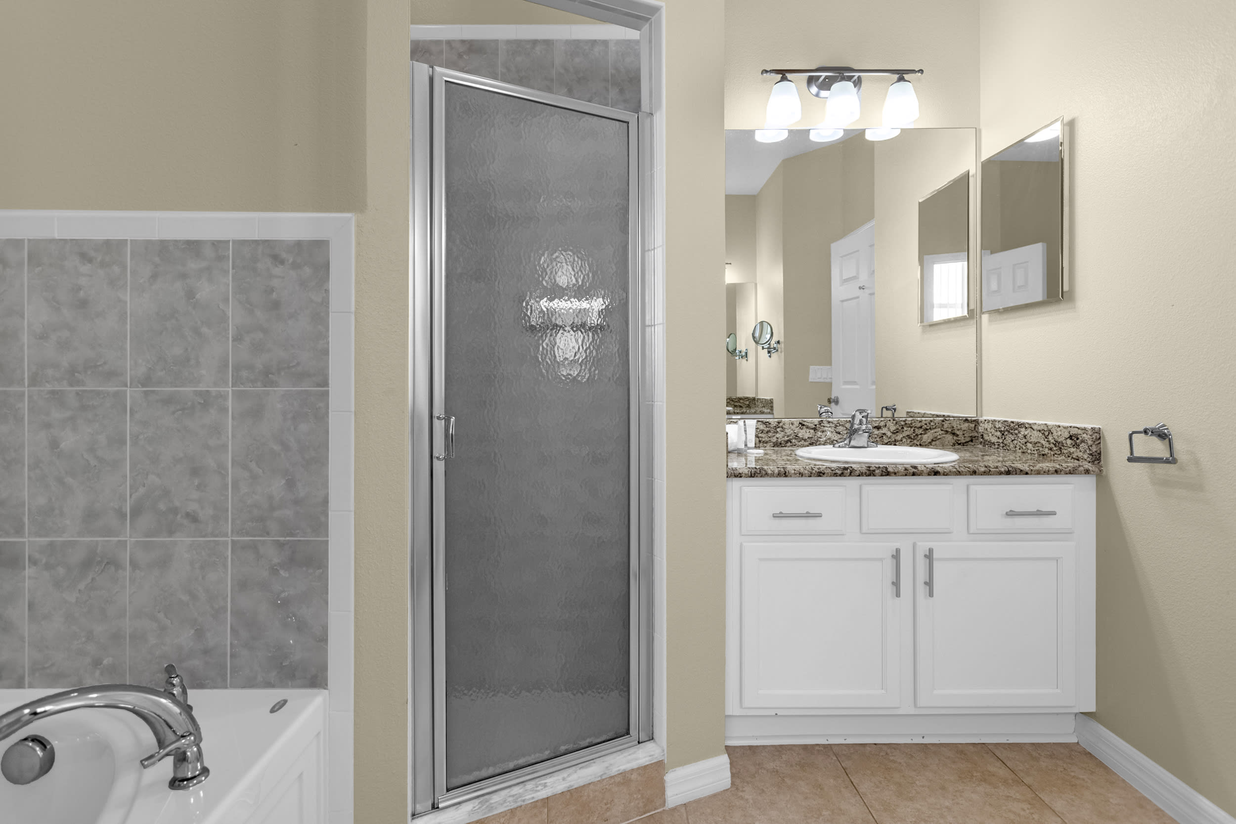 A close-up of the glass-enclosed shower in a bathroom, adjacent to a white built-in bathtub, with a vanity and mirror visible in the reflection, all on light-colored tile floors.
