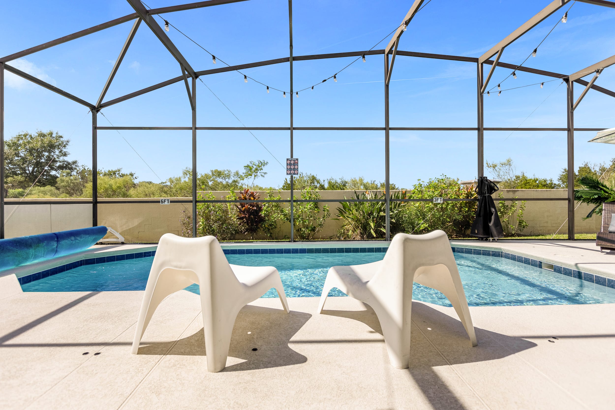 Two modern white lounge chairs are placed on the light-colored patio beside a private screened pool, offering a relaxing spot to enjoy the sunny outdoor environment. The pool features a shallow entry, and string lights are strung across the enclosure.