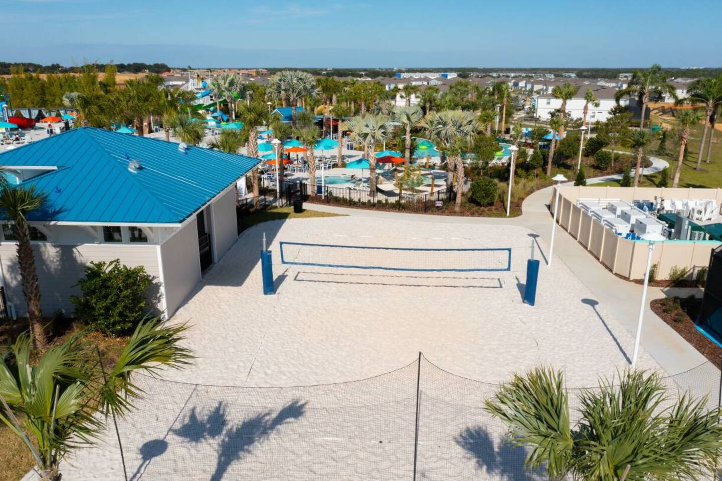 Aerial shot of a resort's outdoor recreation area featuring a pristine sand volleyball court with a blue net in the foreground. Beyond it, a building with a bright blue roof overlooks a sprawling water park with multiple pools, water slides, and numerous palm trees under a clear blue sky.