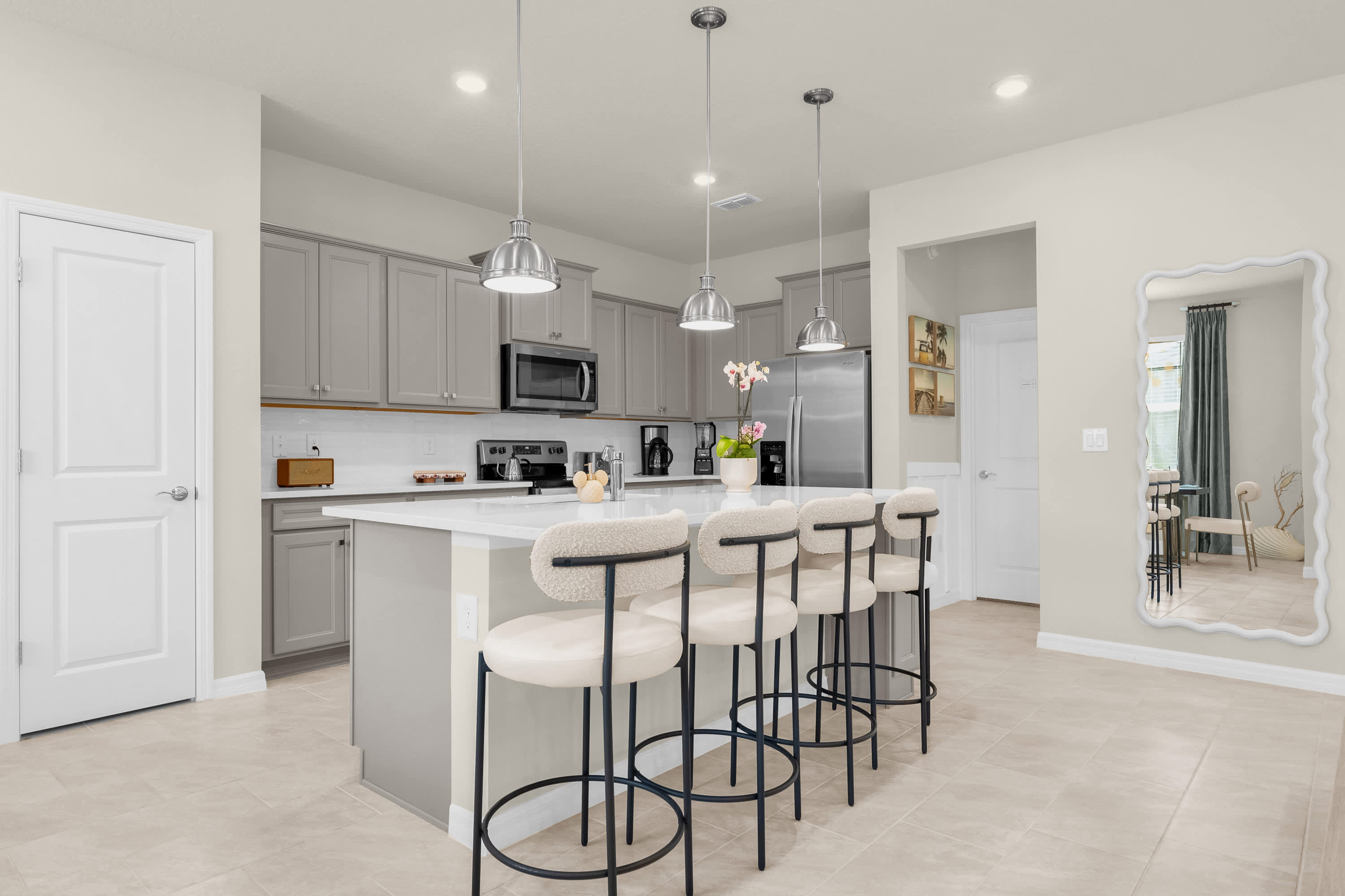 A bright and modern kitchen featuring a large island with a sink and four bar stools, surrounded by light gray shaker cabinets and white countertops. Stainless steel appliances are integrated, and three pendant lights illuminate the island.