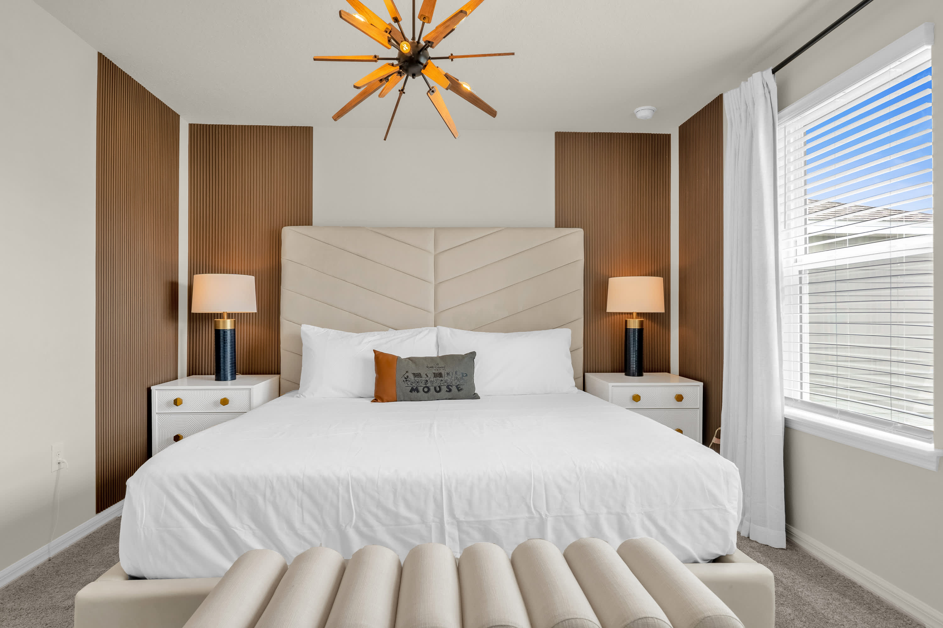 A spacious bedroom showcases a king-sized bed with a cream upholstered headboard, flanked by white nightstands and modern lamps. The room features a unique sputnik-style chandelier and vertical wood paneling accent walls.