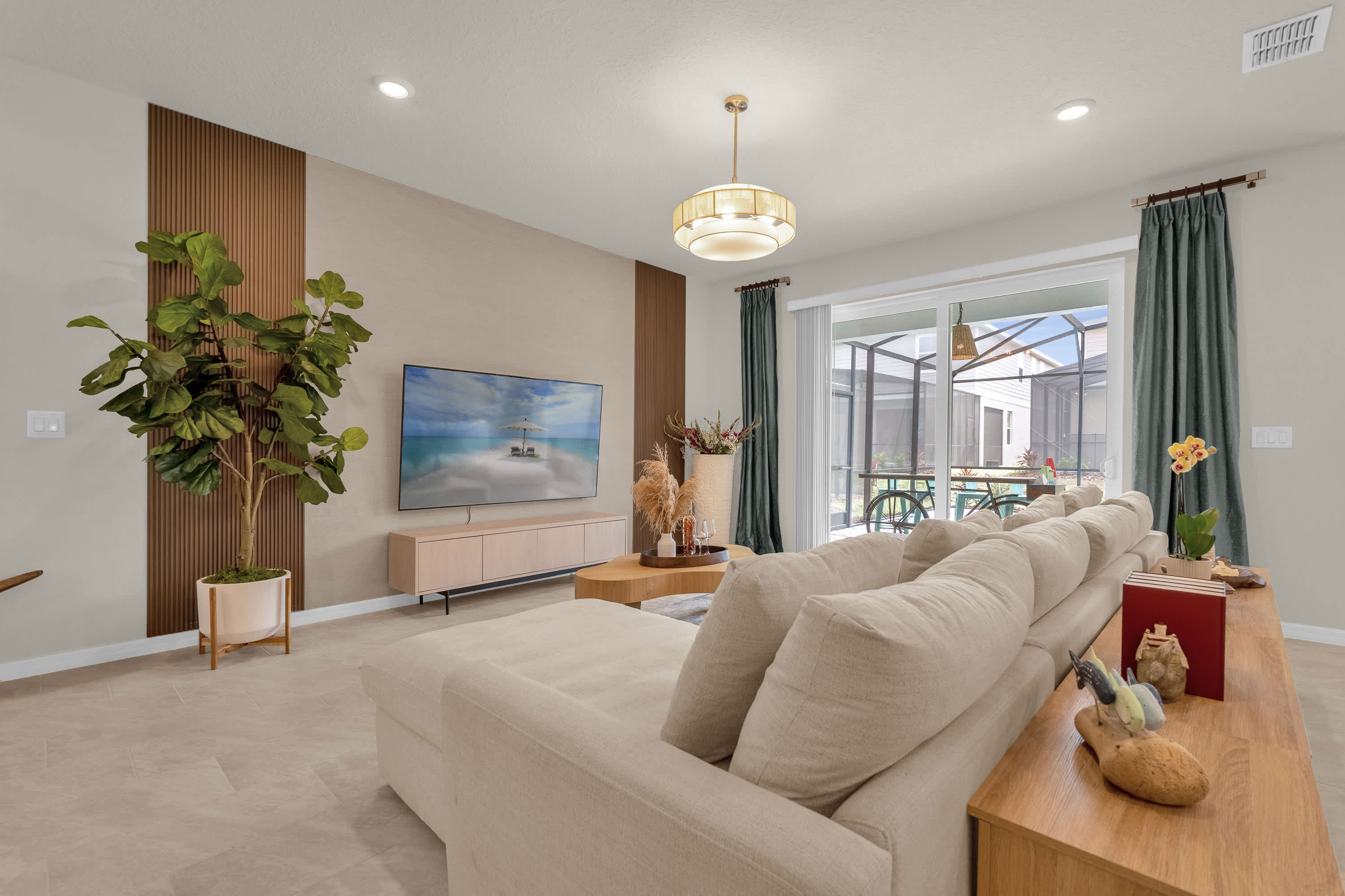 A comfortable living room with a large cream sectional sofa facing a wall-mounted TV and a light wood console. Sliding glass doors with teal curtains open to a screened outdoor area, and a modern pendant light illuminates the space.