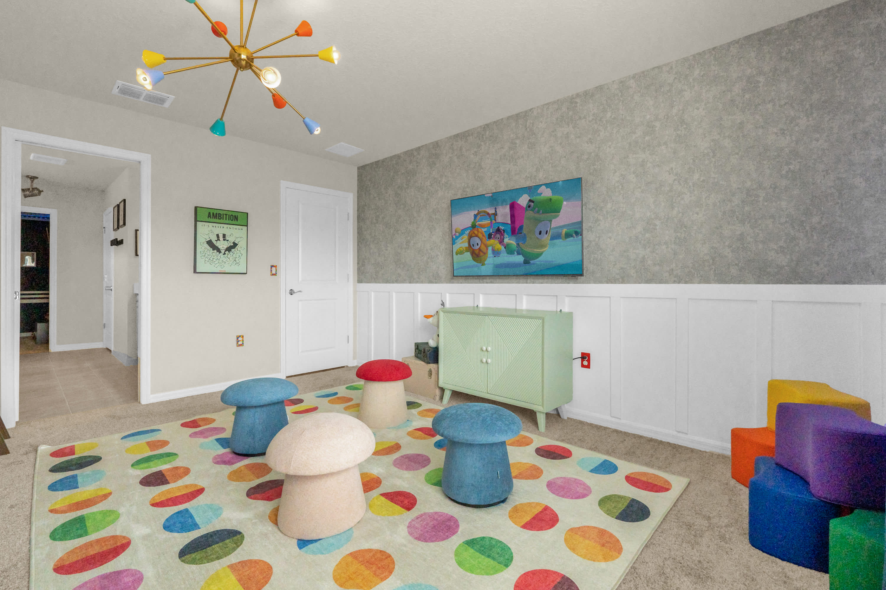 A vibrant kids' play area features a large colorful polka-dot rug, mushroom-shaped seating, and soft play blocks, set against a wainscoted wall with a mounted TV. A unique sputnik-style chandelier hangs from the ceiling.