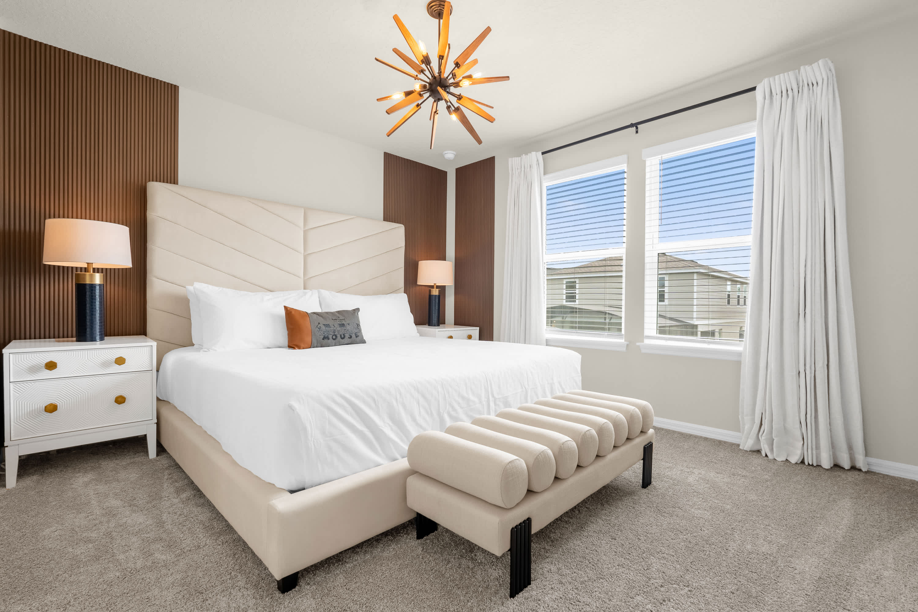 A bright bedroom features a king-sized bed with a tall upholstered headboard, flanked by white nightstands with gold accents and modern lamps. A striking sputnik-style chandelier hangs from the ceiling, complementing the accent wall paneling.