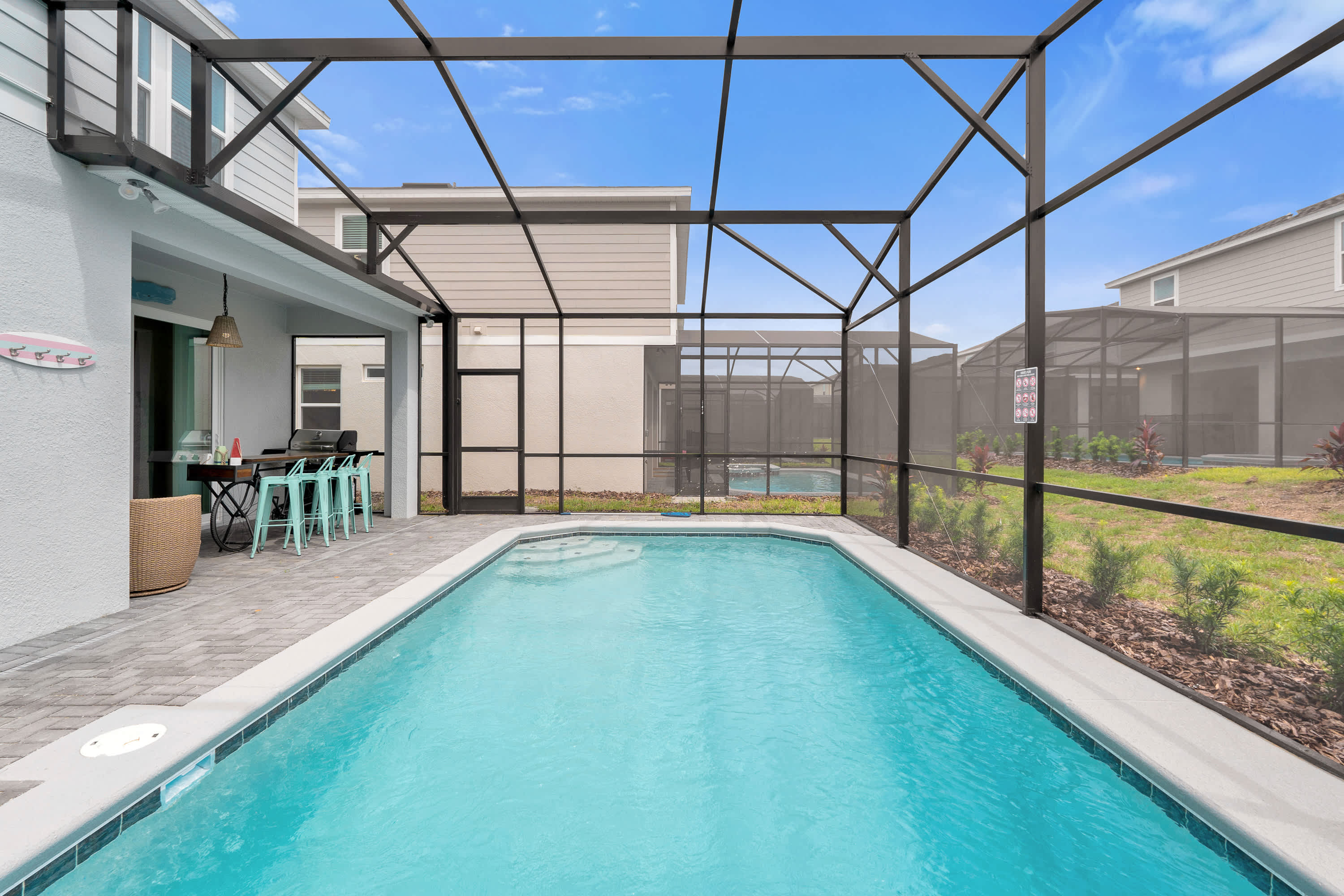 A private rectangular swimming pool within a screened enclosure, surrounded by a paved patio and offering a view of the adjacent houses and a small grassy area.