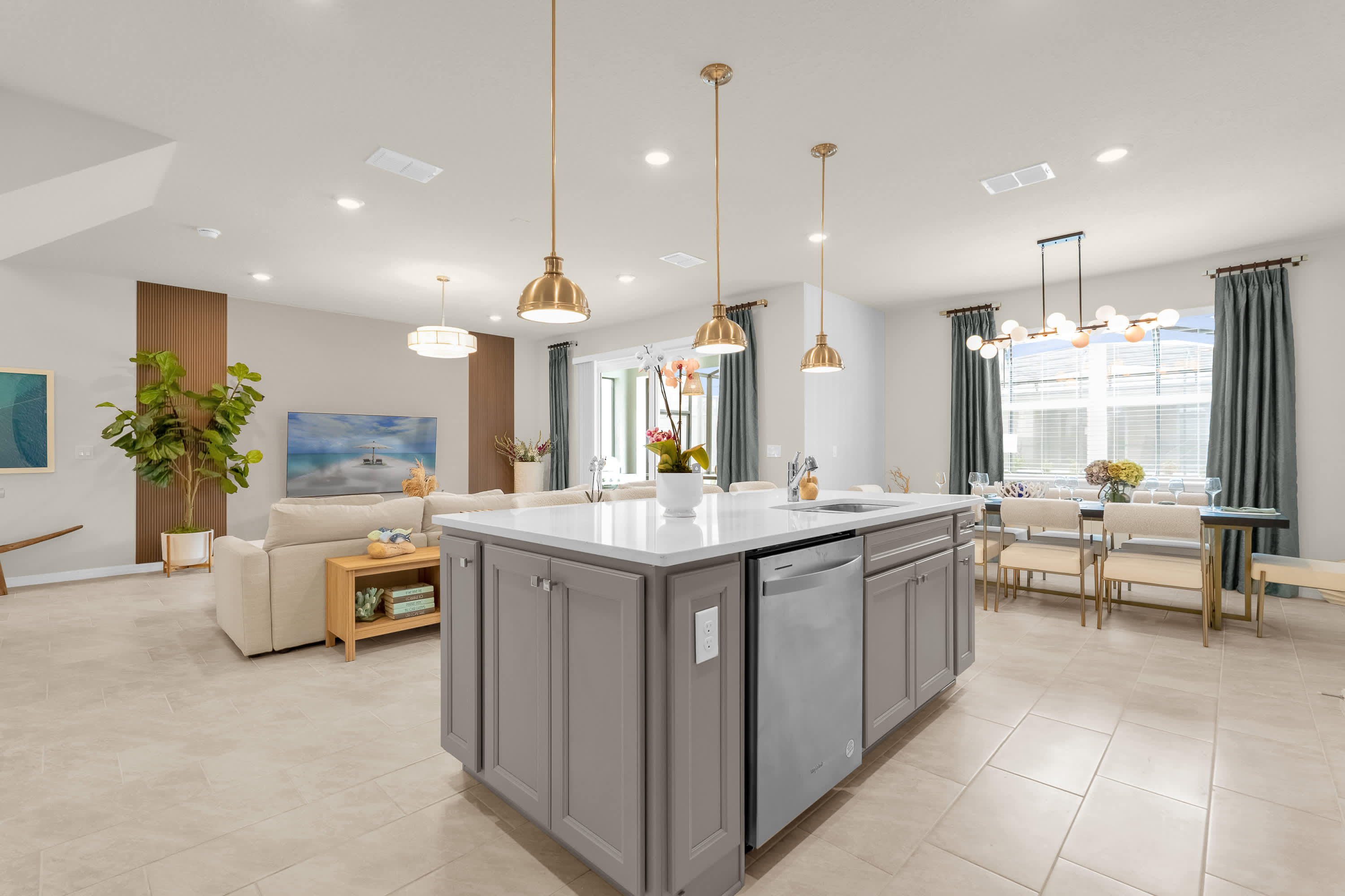 Modern open-concept kitchen featuring a large gray island with white quartz countertops and three brass pendant lights, overlooking a dining area and living room with light-colored tile flooring.