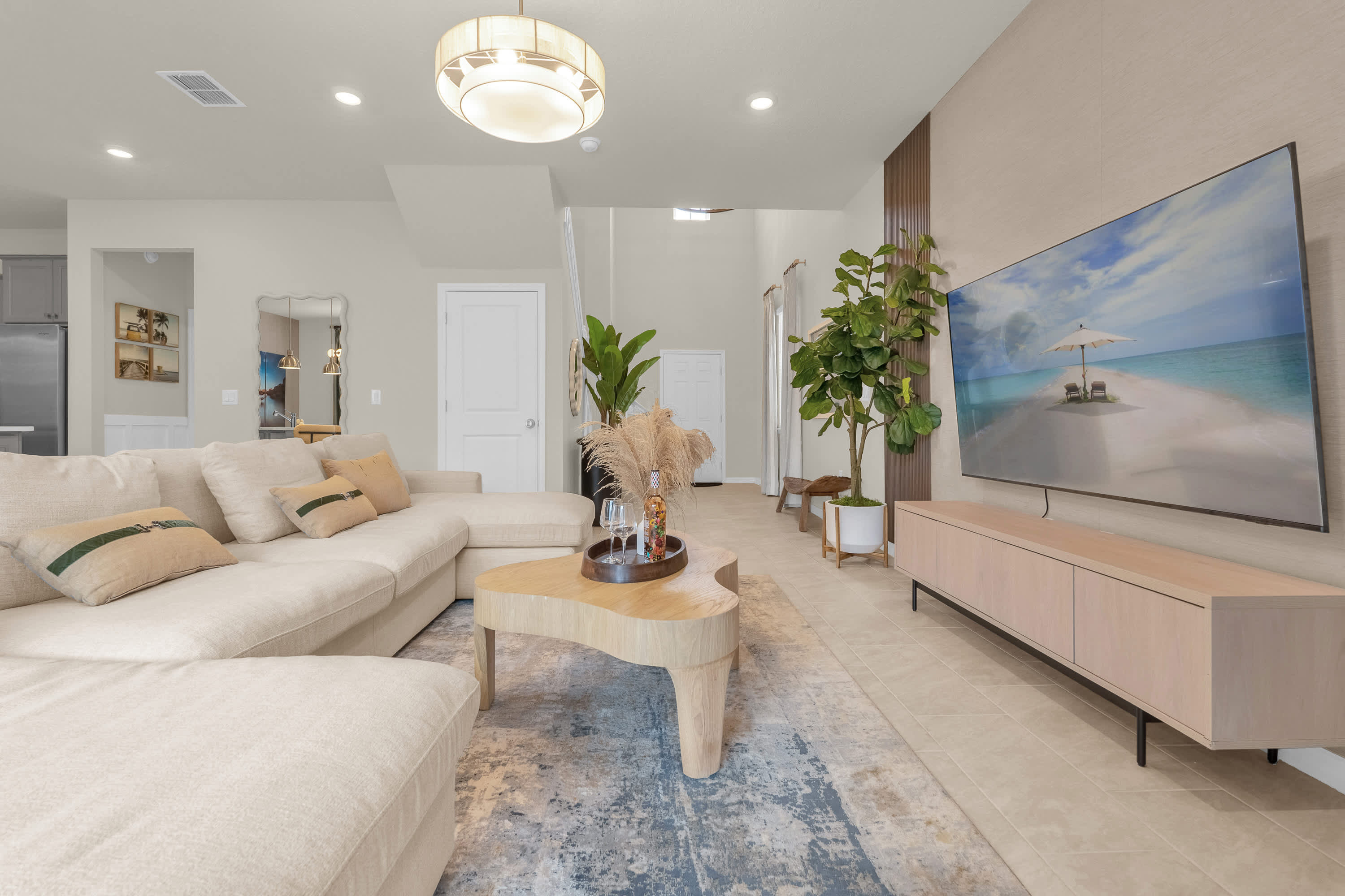 A spacious living room featuring a large cream-colored sectional sofa with throw pillows, centered around a modern wooden coffee table on a patterned area rug. A large wall-mounted TV sits above a light wood console, flanked by a tall plant.