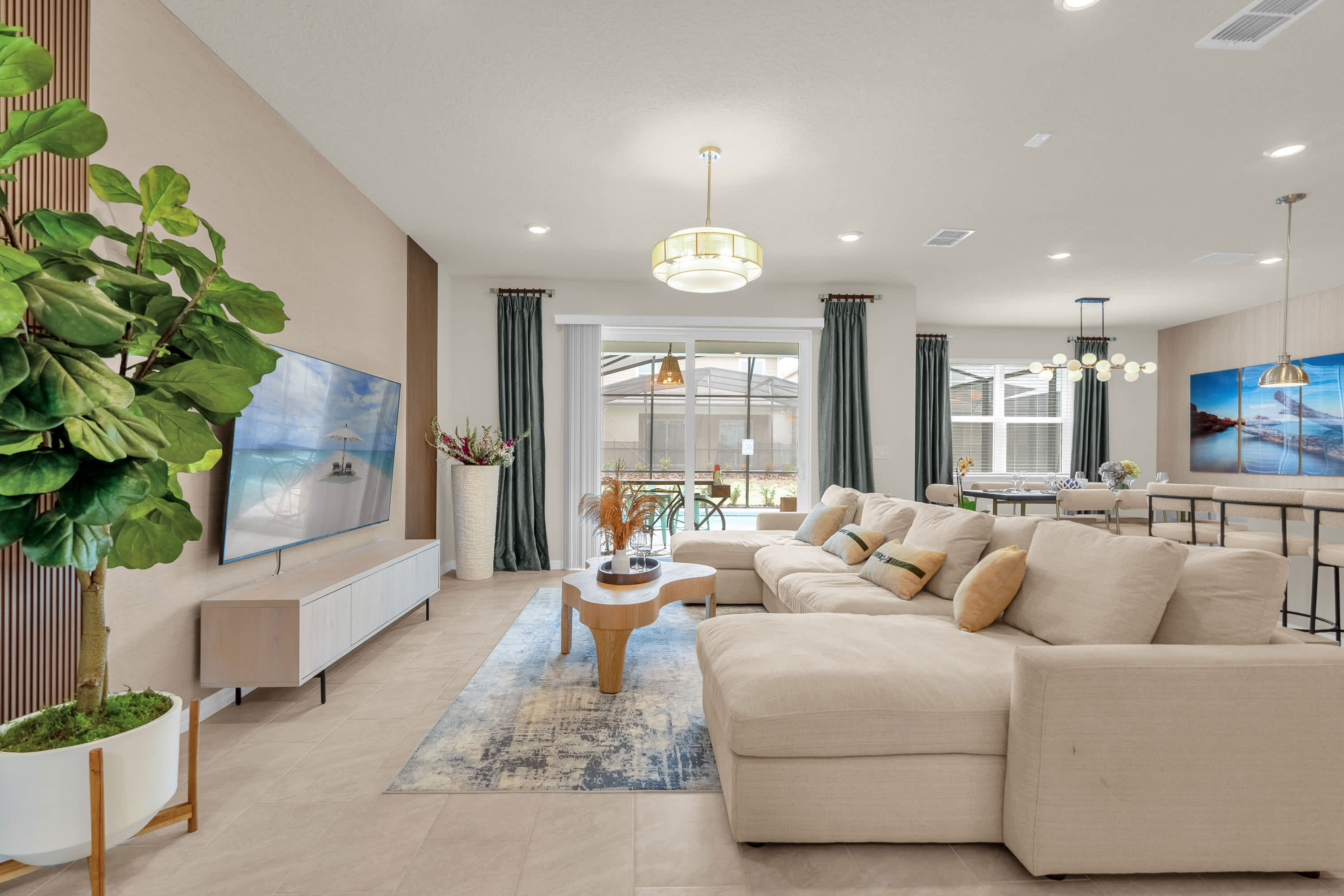 A spacious and bright living room featuring a large L-shaped sectional sofa in a neutral tone, a modern coffee table, and a wall-mounted flat-screen TV. Large sliding glass doors lead to the outdoor pool area.