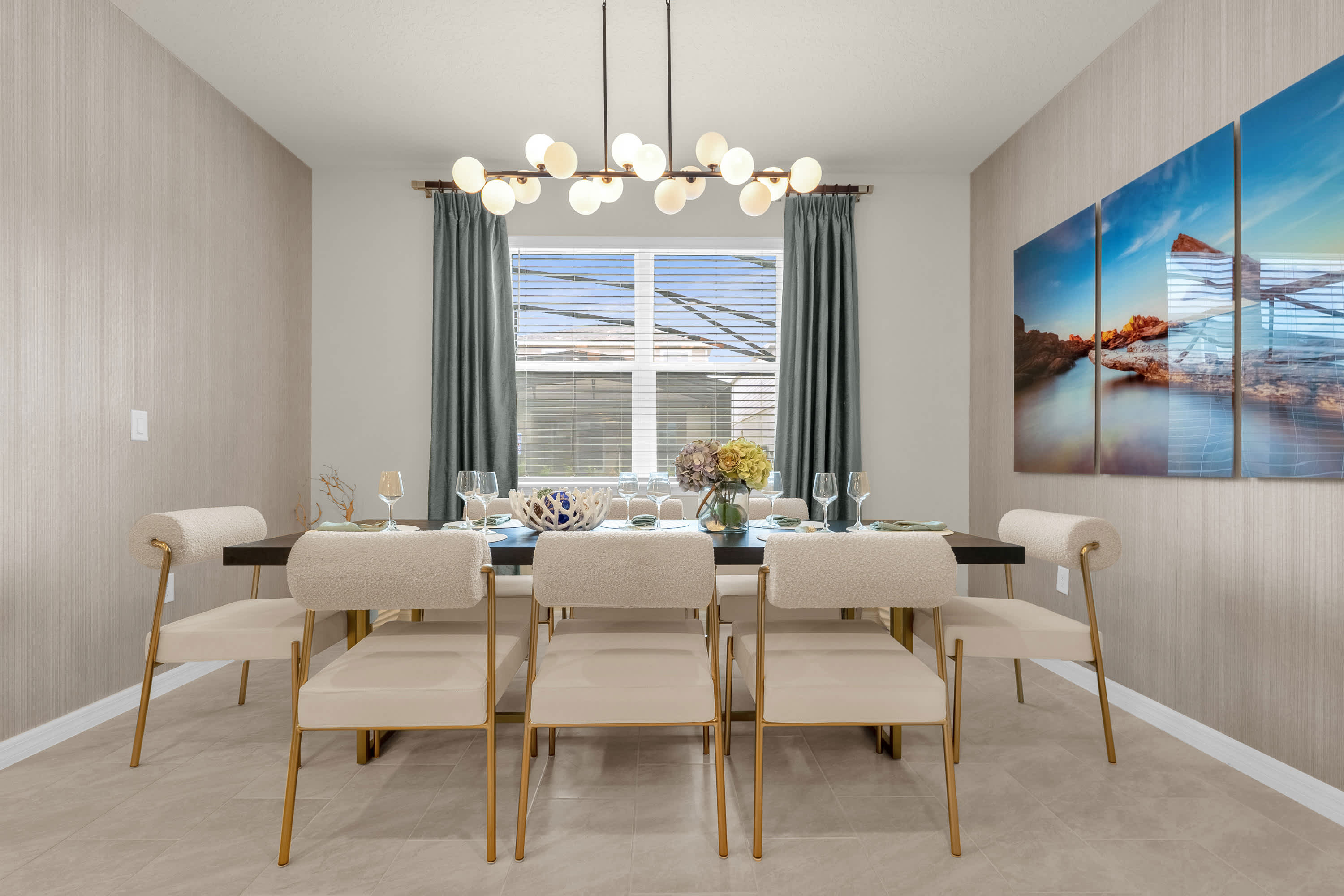 An elegant dining room with a long dark wood table set for multiple guests, surrounded by modern chairs with gold frames and cream upholstery. A contemporary linear chandelier hangs above the table, and large windows provide natural light.