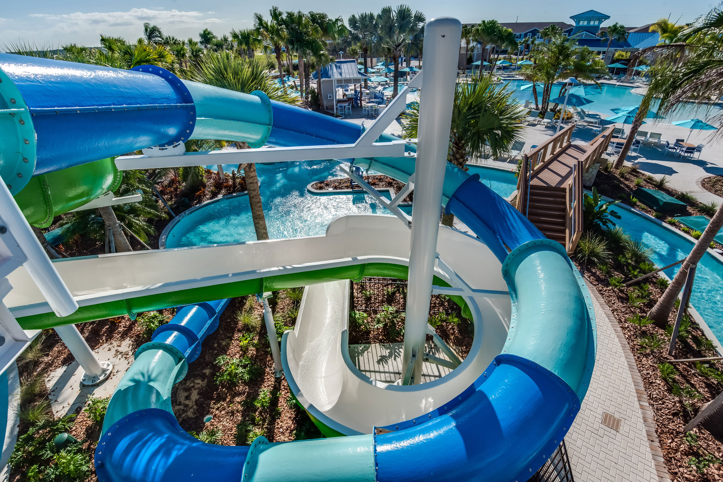 A close-up aerial view of a large, multi-colored water slide structure with blue, green, and white slides twisting down into a pool area. A wooden bridge and a lazy river are visible below.