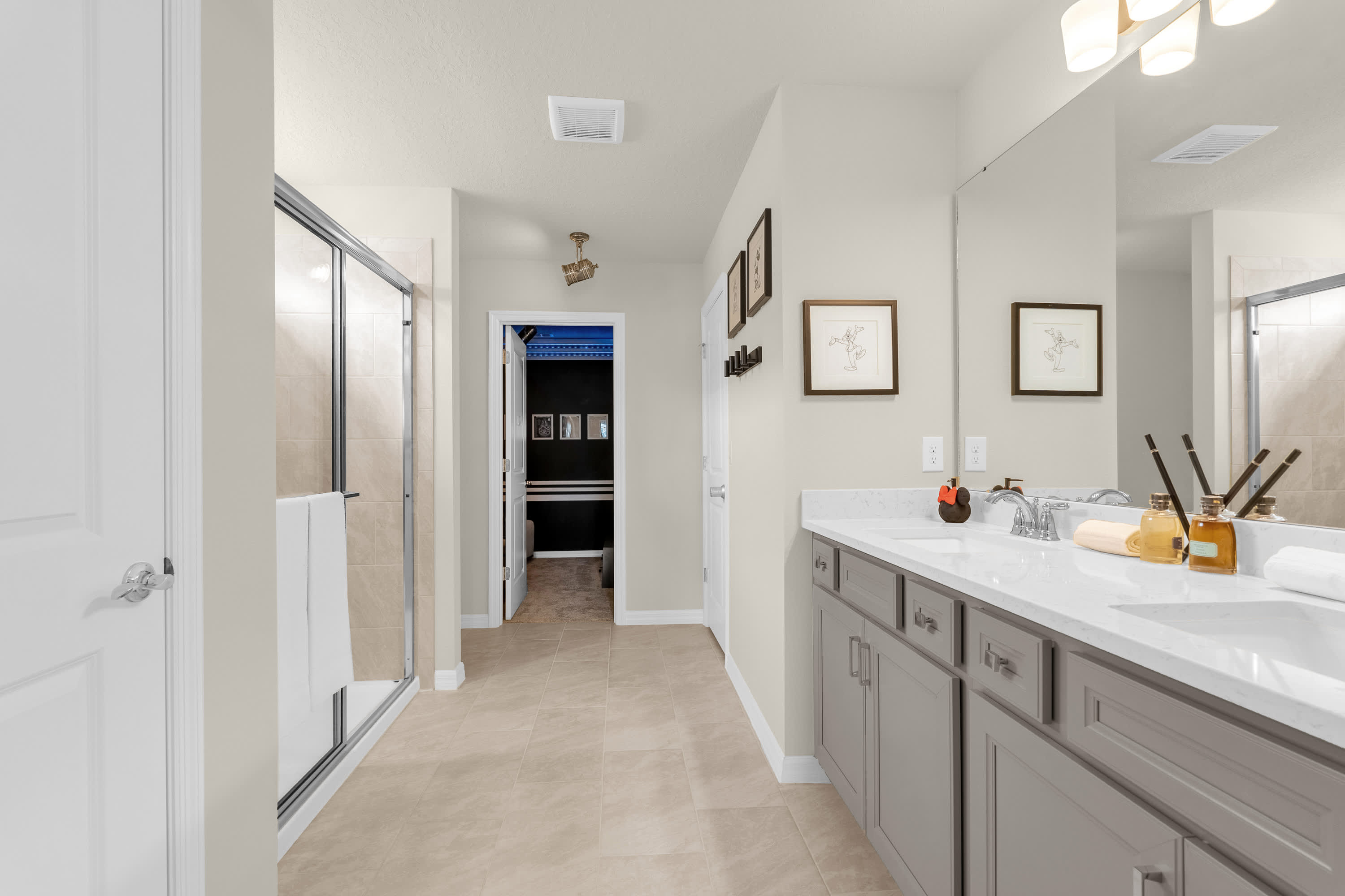 A spacious bathroom features a double vanity with gray cabinets and white countertops, large mirrors, and a walk-in shower with tiled walls. The room has light-colored tiled floors and neutral walls.