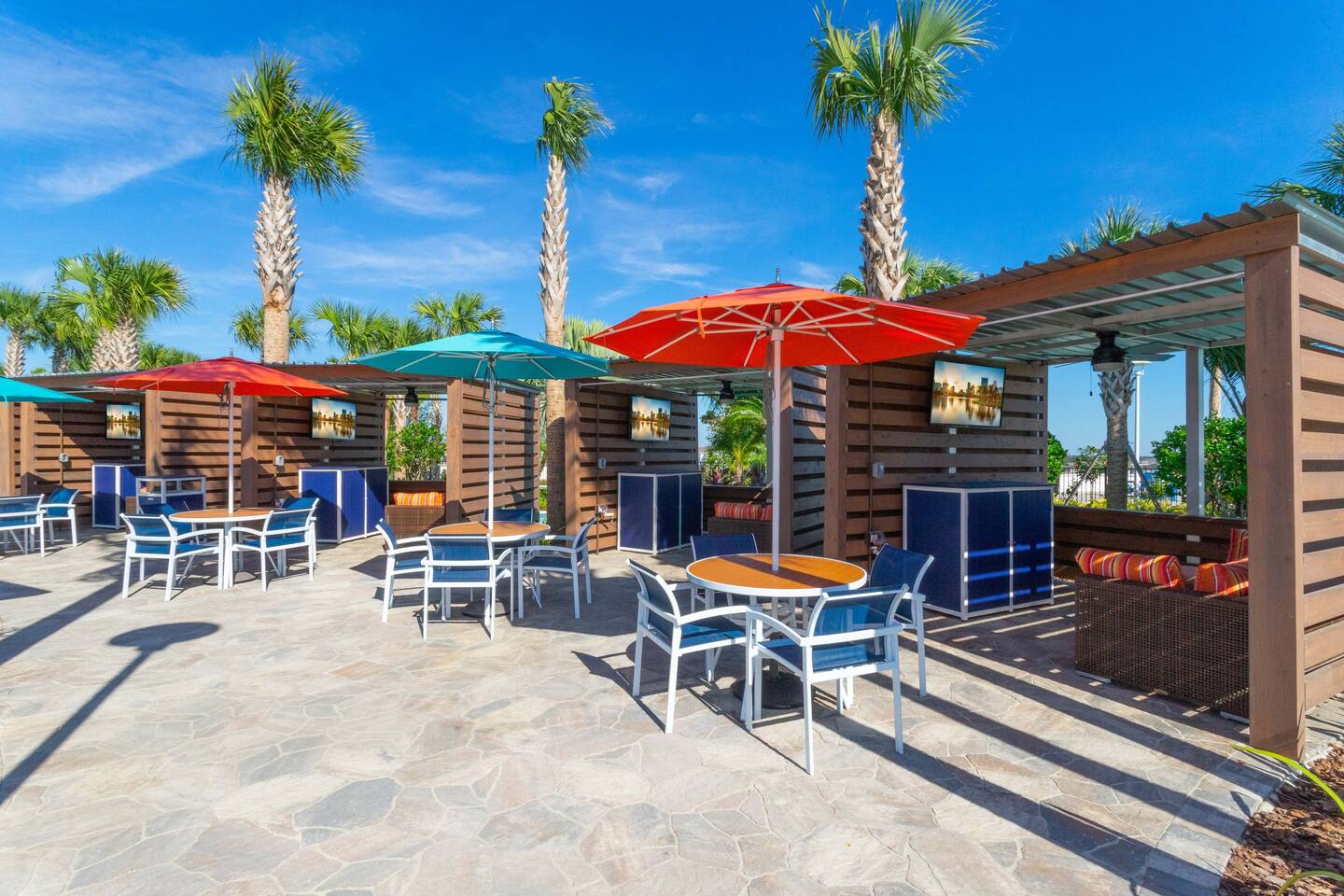 An outdoor seating area features multiple dining tables with chairs and colorful umbrellas (orange, teal), set on a patterned stone patio. Private cabanas with TVs and lounge seating are visible in the background.