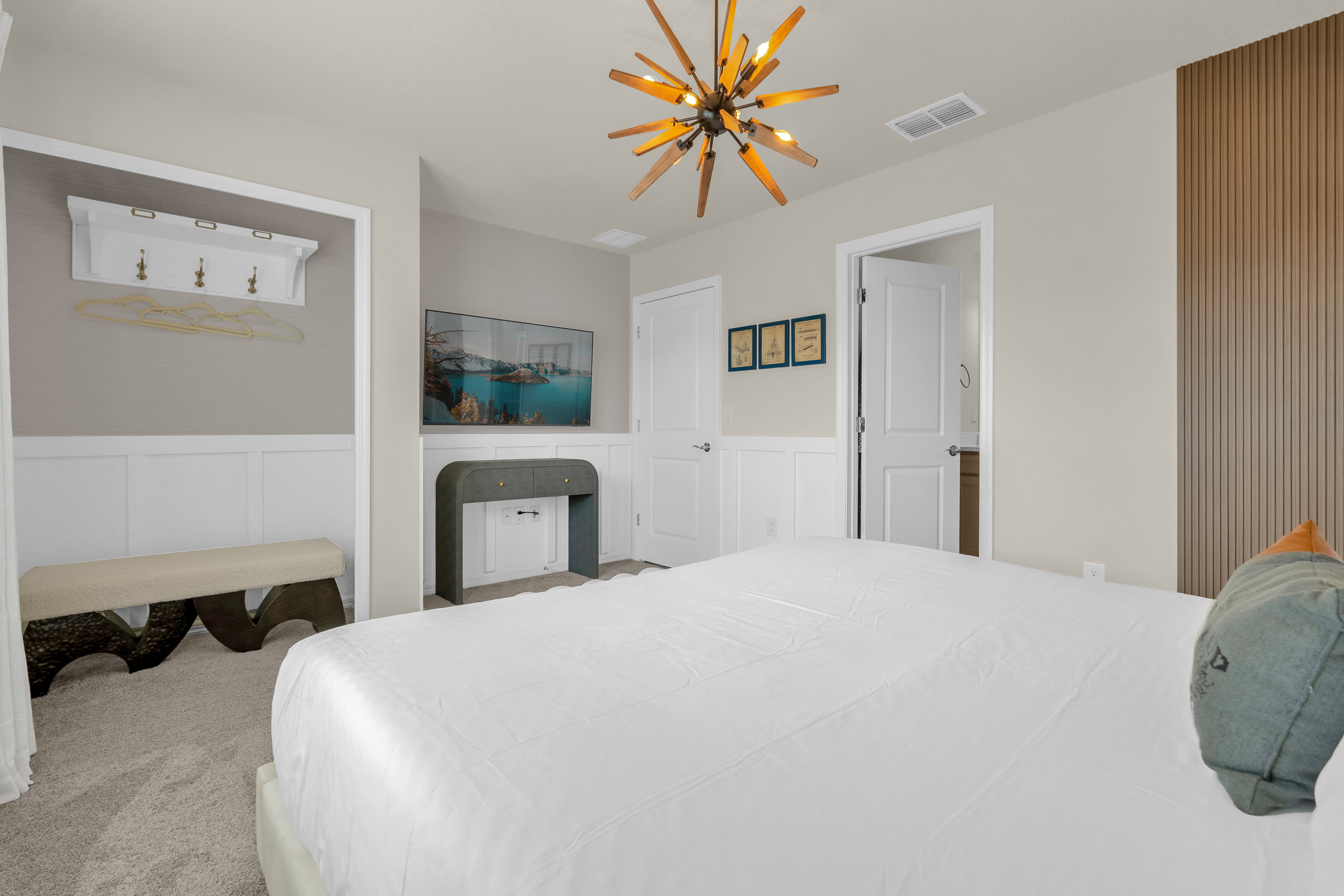 This view of the bedroom highlights a wall-mounted TV above a console, a long upholstered bench, and a small open closet area with a coat rack. White wainscoting and light walls create a clean aesthetic.