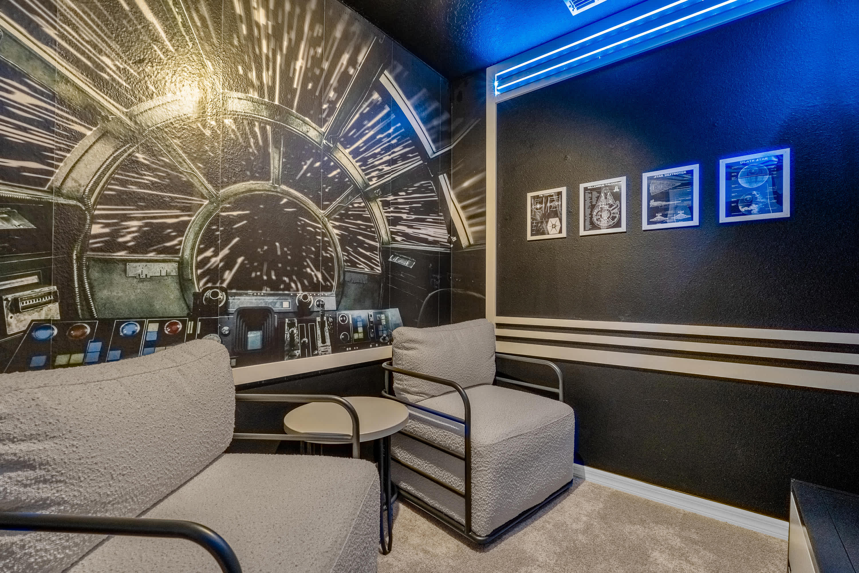 A dark, themed media room features a large Star Wars Millennium Falcon cockpit mural on one wall and two cozy gray armchairs with a small side table. Blue LED strip lighting illuminates the ceiling, enhancing the cinematic atmosphere.