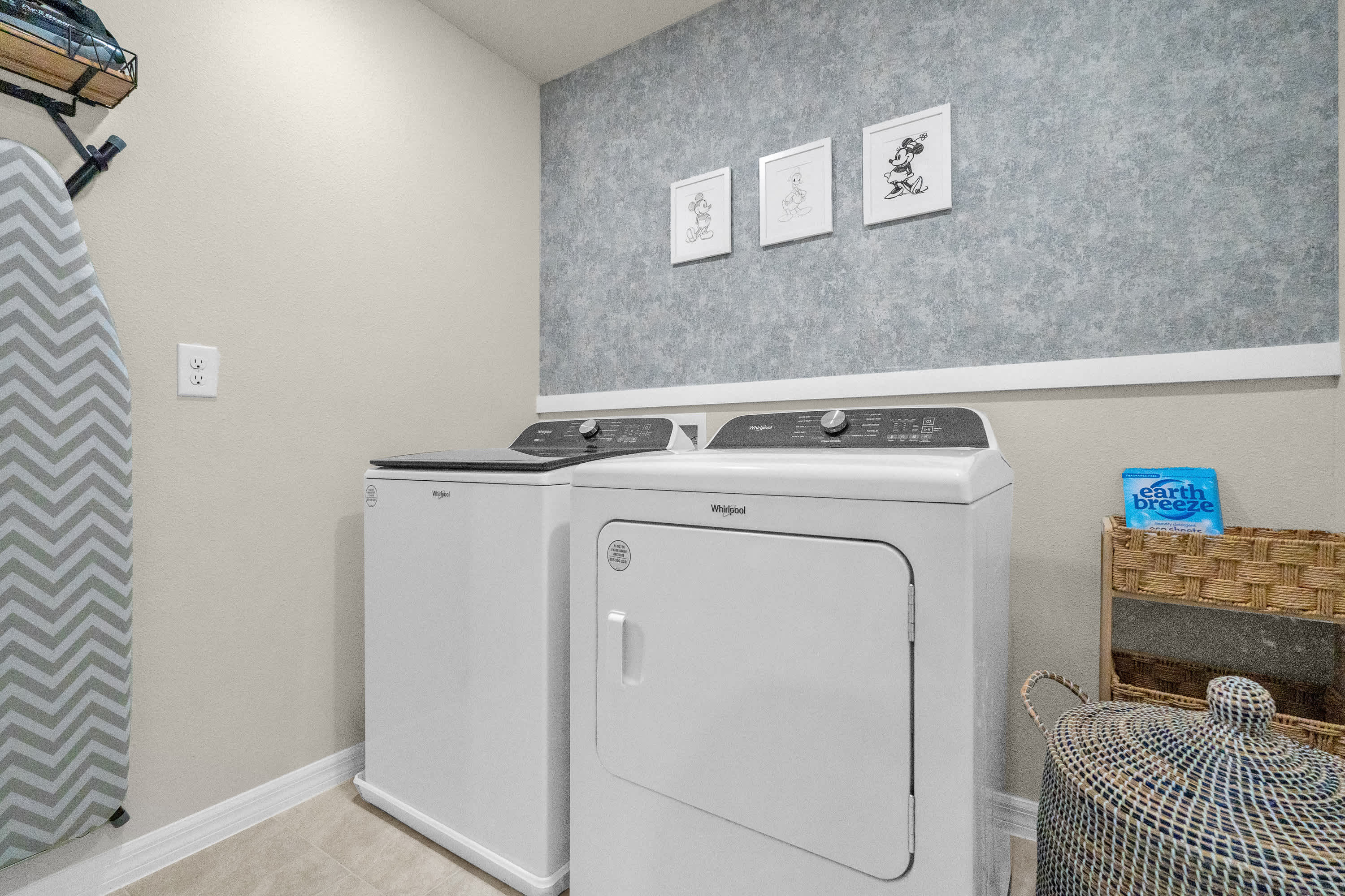 A bright laundry room features a white Whirlpool washer and dryer set, with light beige walls and a textured blue accent wall adorned with three framed Mickey Mouse art prints. A wicker basket and an ironing board are visible on the left.