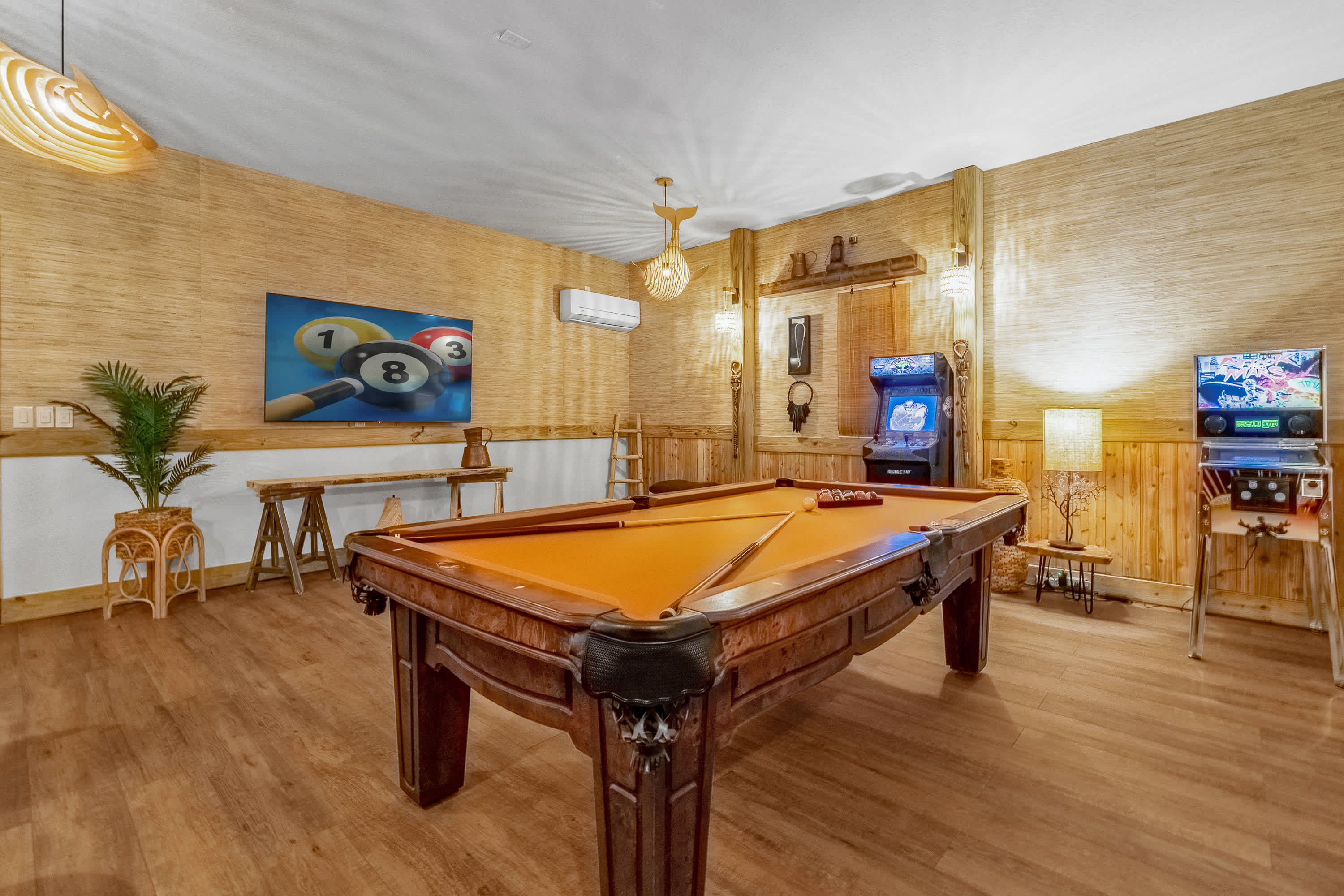 A game room featuring a large wooden pool table with orange felt, positioned on light wood flooring. The walls are a mix of light wood paneling and woven material, with two arcade machines and a large framed artwork of pool balls visible.