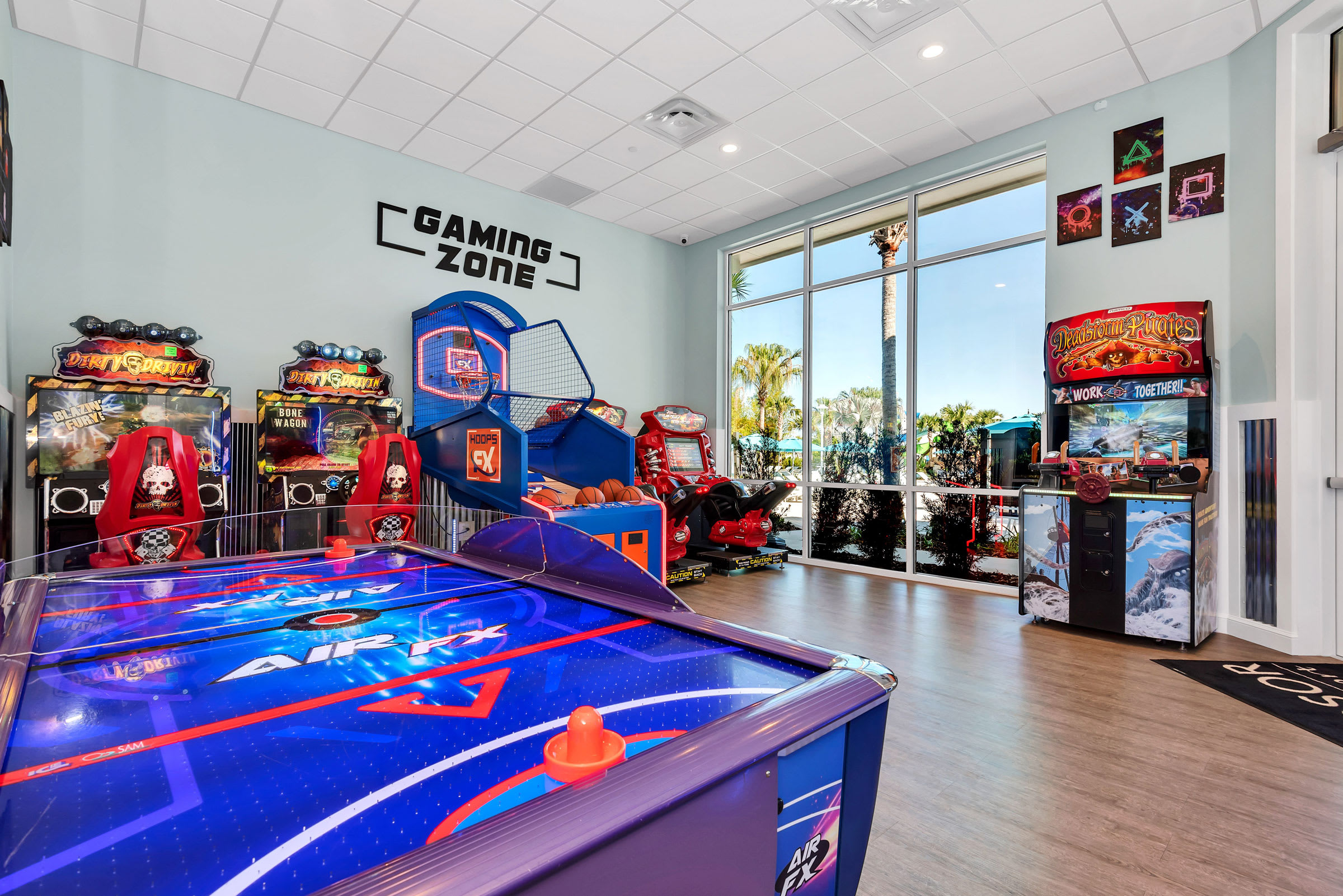 Vibrant indoor game room filled with arcade machines, including a prominent air hockey table in the foreground, a basketball shooting game, and various video game cabinets. Large windows provide a glimpse of outdoor palm trees, and the room features light wood-look flooring and a 'GAMING ZONE' sign on the wall.