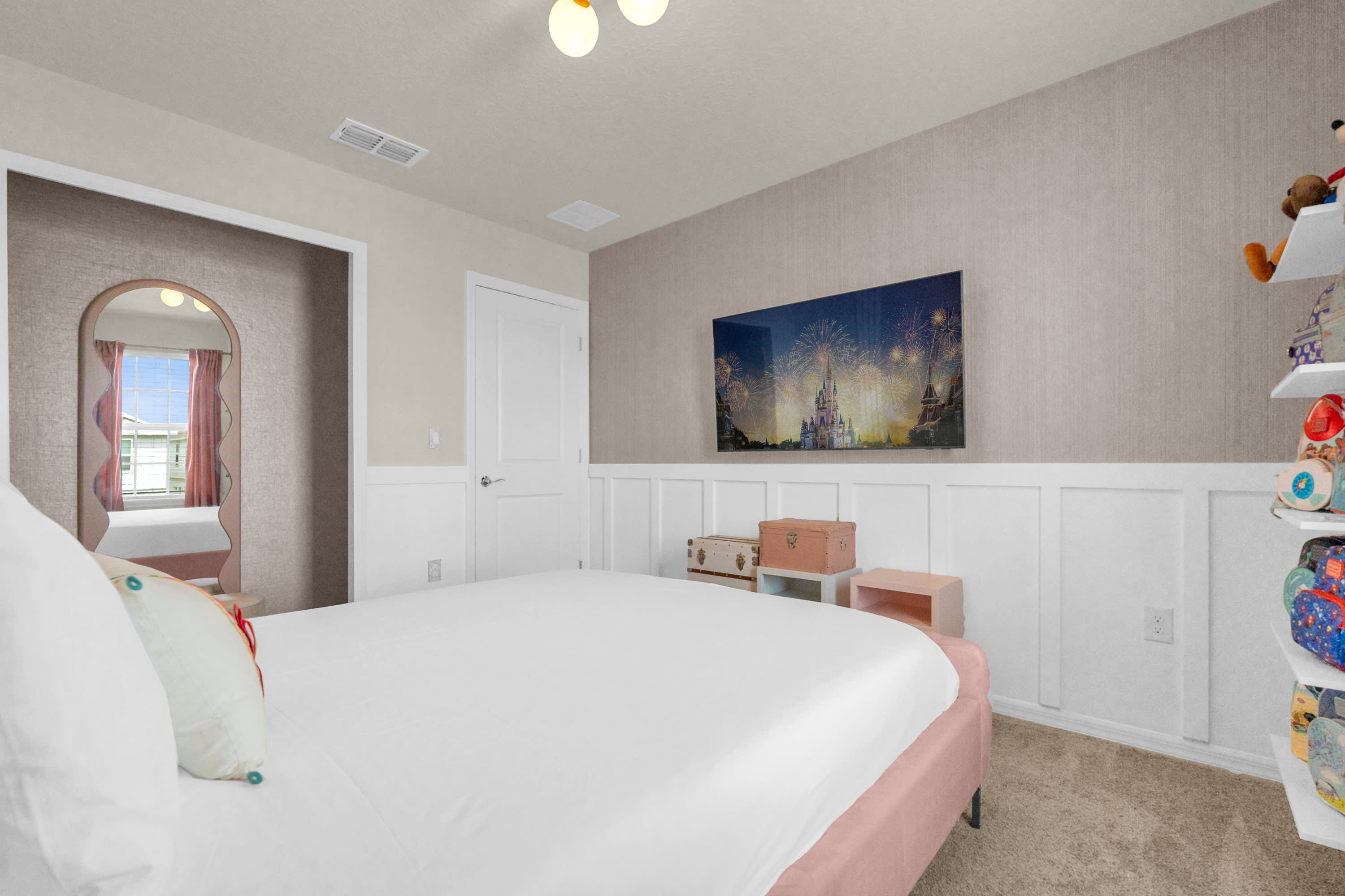 View of the pink-themed bedroom showing a wall-mounted television, a white wainscoting detail along the lower wall, and a vintage-style trunk, contributing to the room's charming decor.