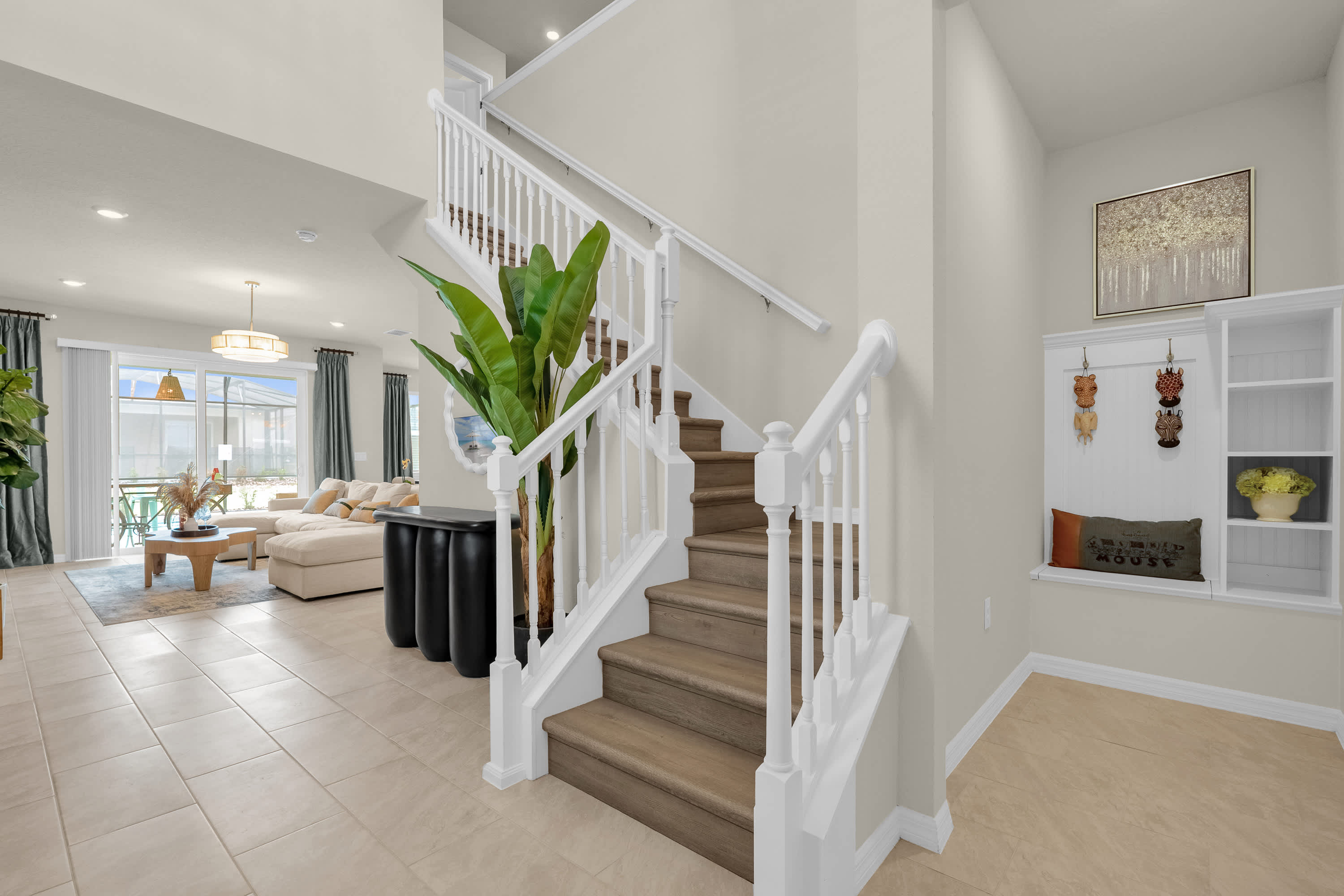 Bright entryway featuring a wooden staircase with white banisters, a large green plant, and a built-in bench with shelving and decorative items, all set on light-colored tile flooring.
