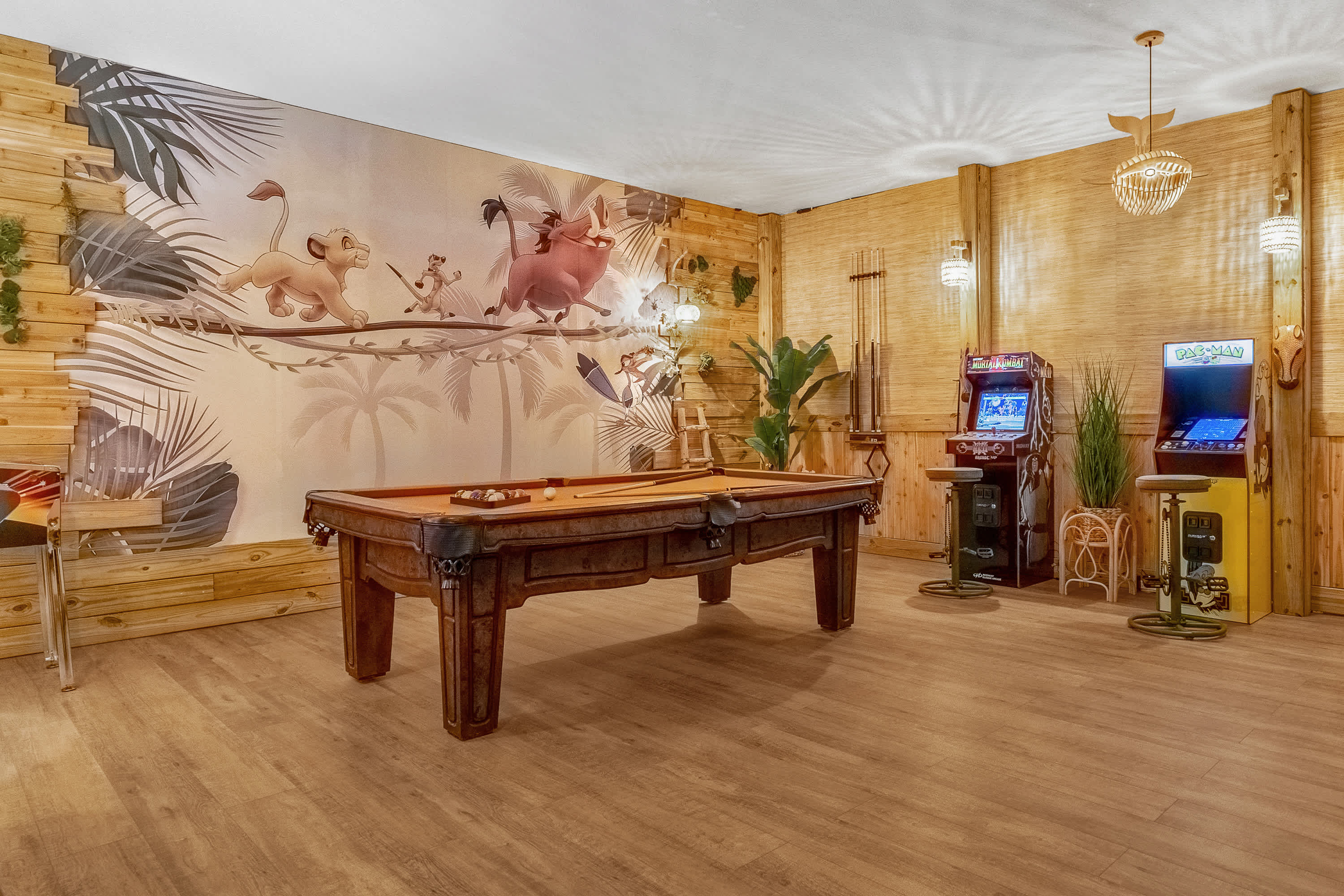 A vibrant game room featuring a wooden pool table with green felt, set against a wall adorned with a large Lion King mural. Two arcade machines stand to the right, and the room is illuminated by recessed and decorative pendant lighting.