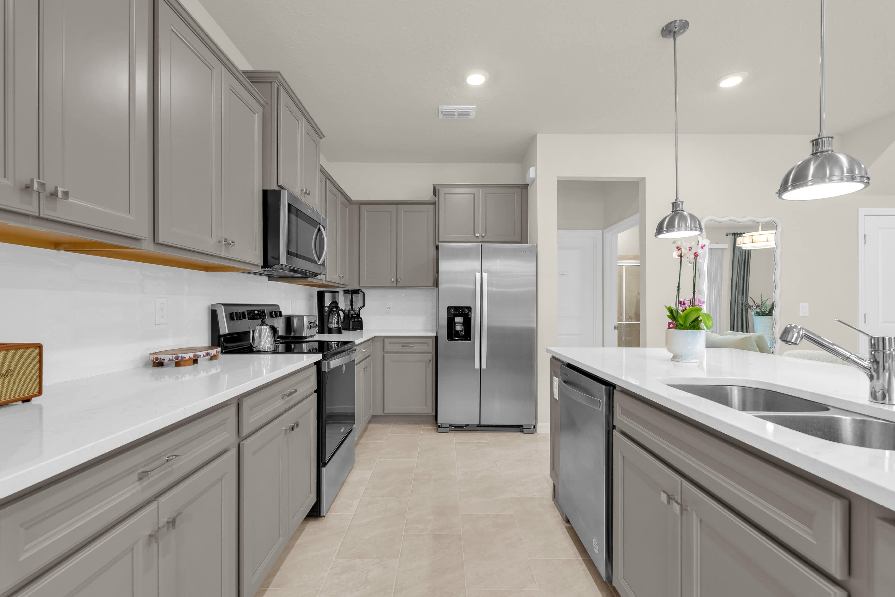 A well-equipped galley-style kitchen with light gray shaker cabinets, white countertops, and stainless steel appliances including a refrigerator, microwave, and stove. The light tile flooring extends throughout the space, offering a clean and functional design.