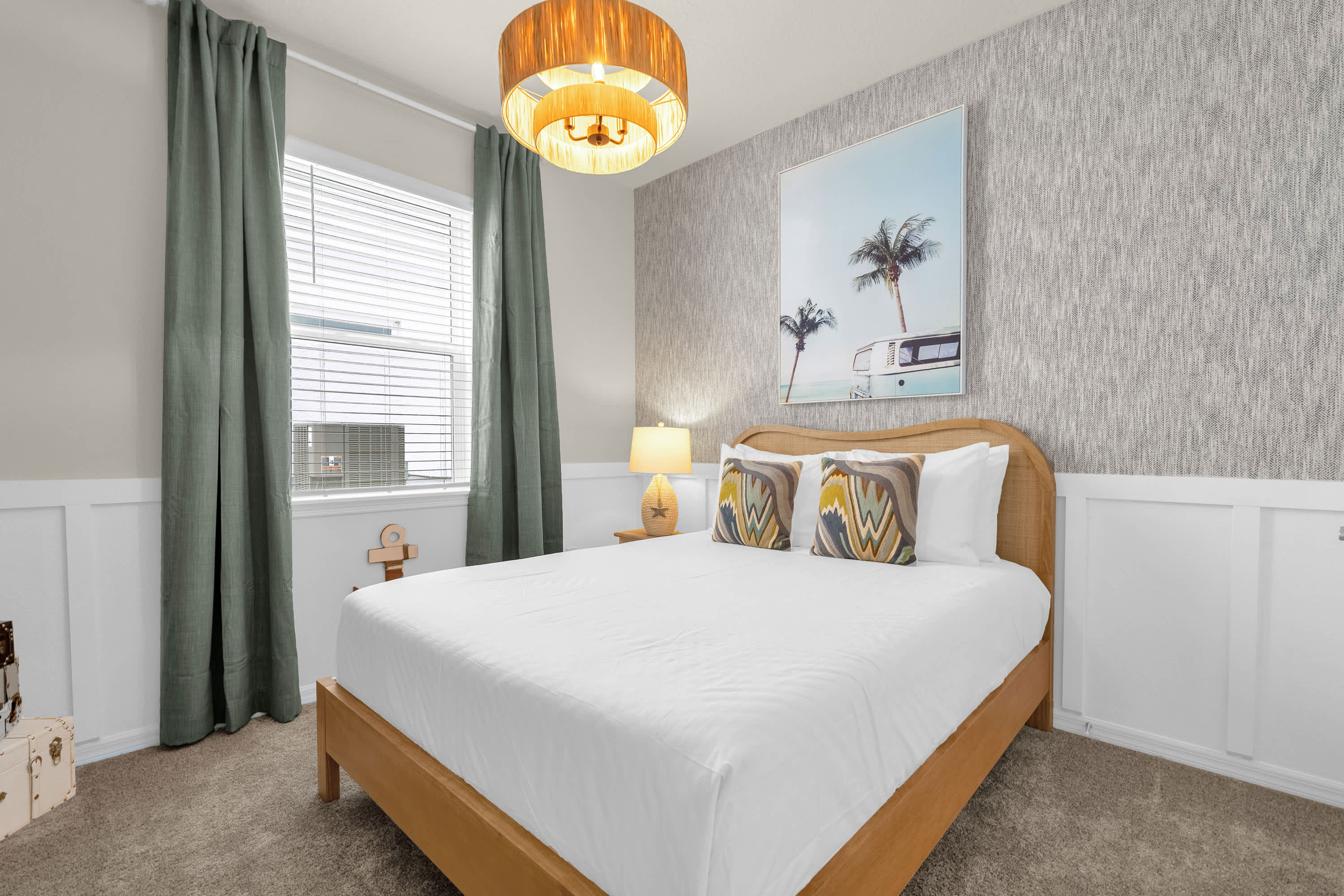 A cozy bedroom features a queen-sized bed with a wooden headboard, flanked by a nightstand with a coastal-themed lamp. The room has wainscoting, a textured accent wall, and a distinctive drum-style ceiling light.